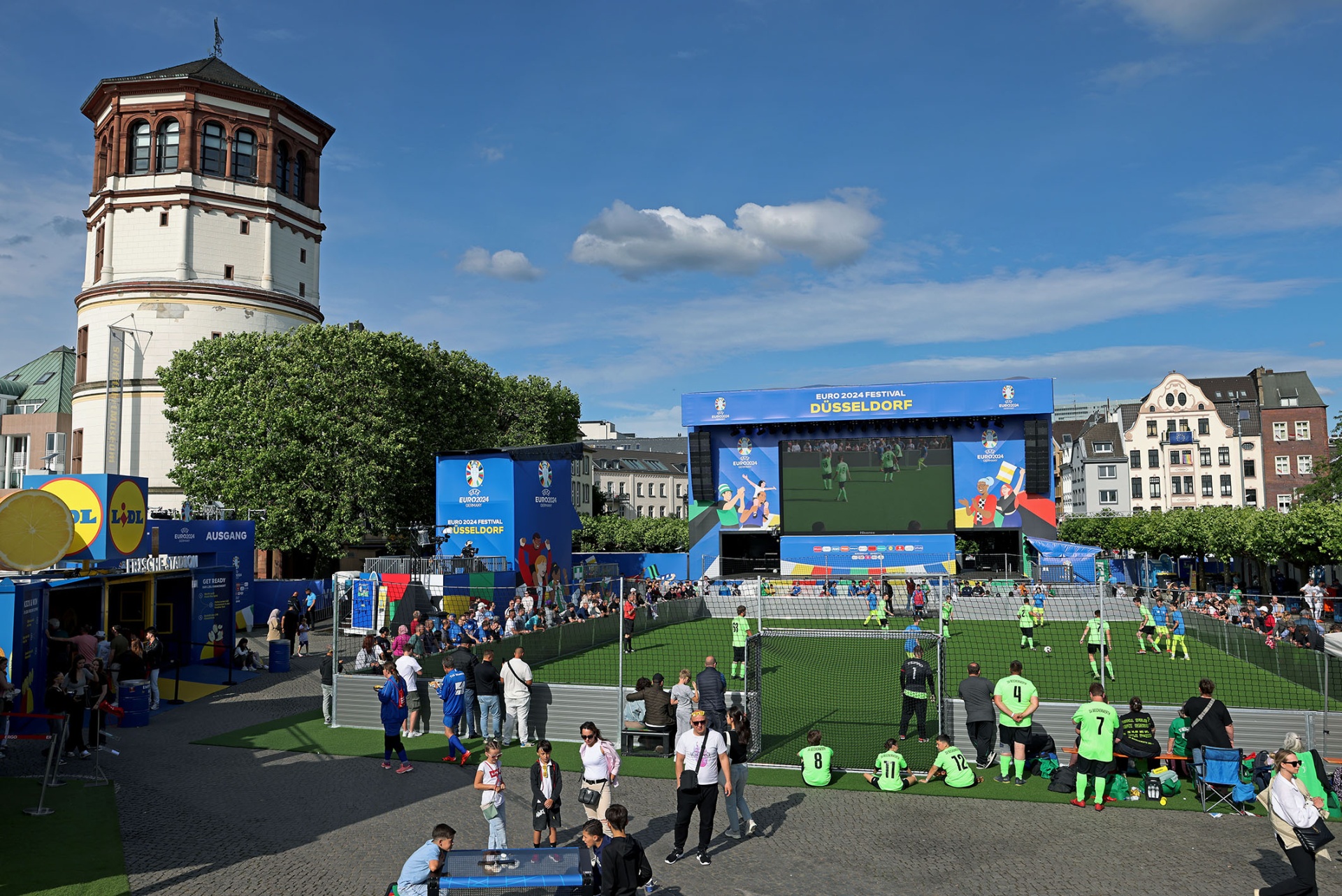 [{"clang_id":1,"value":"Düsseldorfer Platz: Fußballspiel auf Rasen vor Bühne mit Großbildschirm. Im Hintergrund der Rheinturm und Gebäude. Menschengruppen, Zelte, sonniger Tag."}]