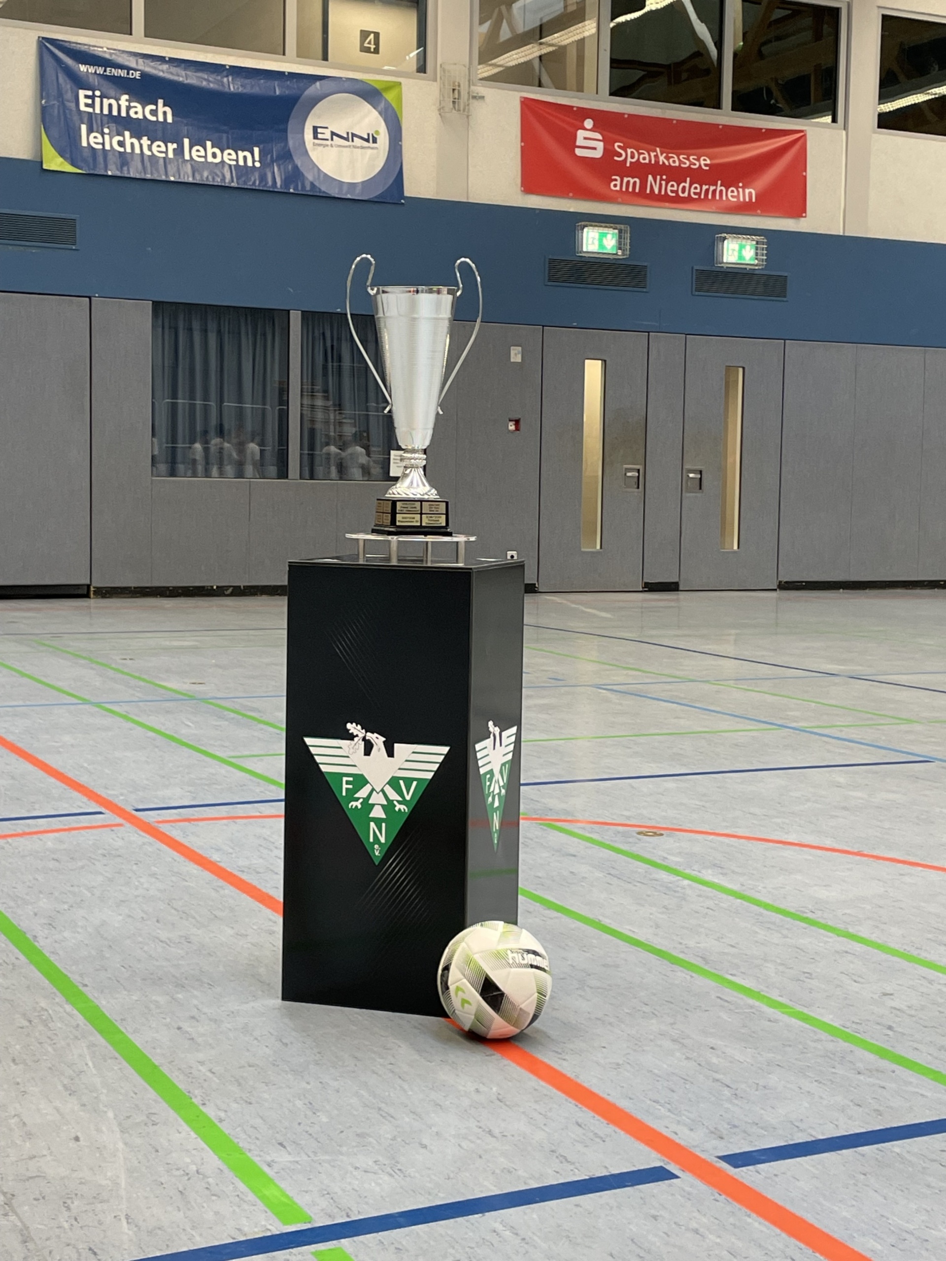 [{"clang_id":1,"value":"Fußballtrophäe auf grün-schwarzem Podest in Sporthalle. Davor Fußball. Im Hintergrund Banner und Fenster. Farbige Markierungen auf dem Hallenboden."}]