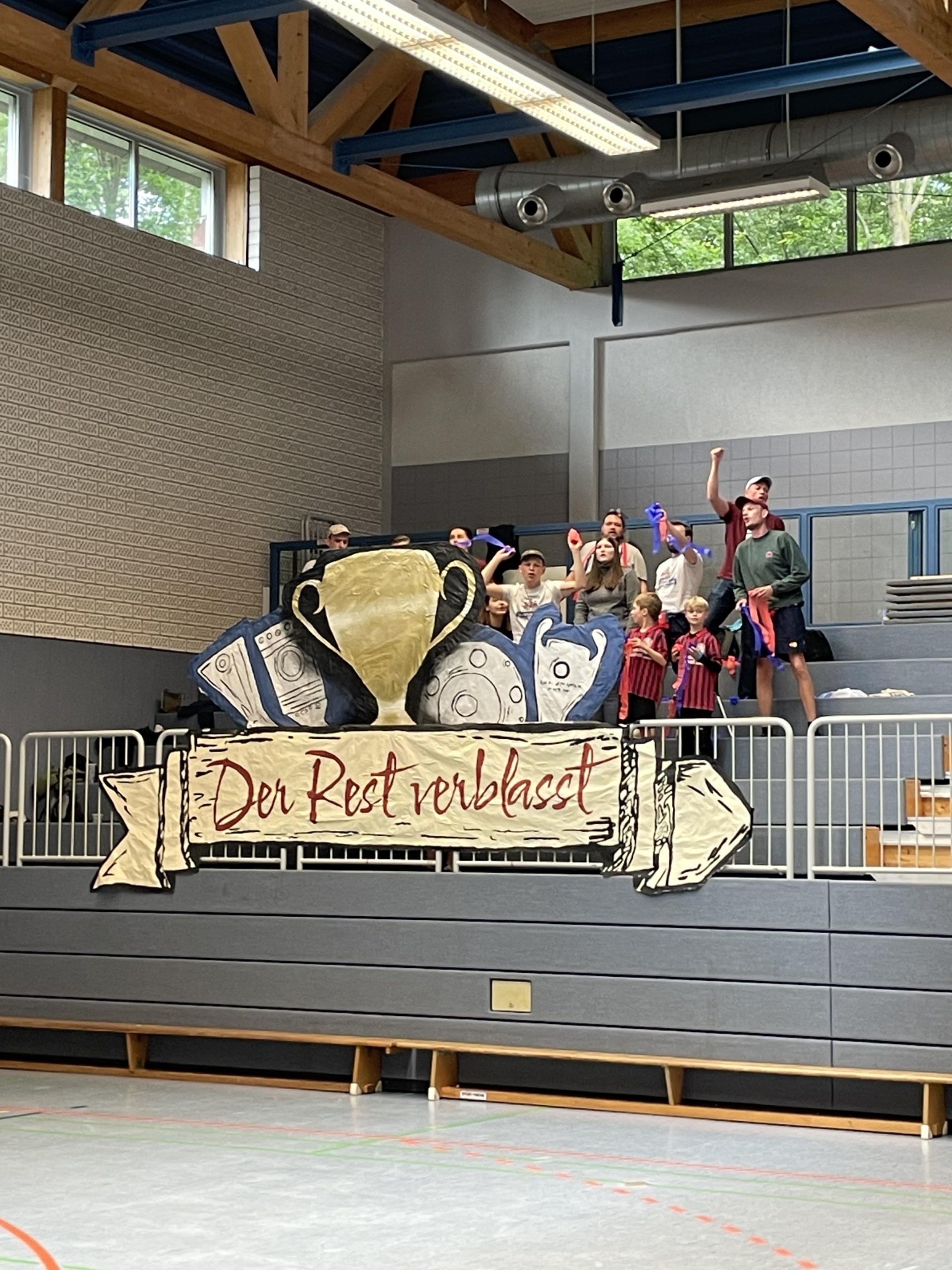 [{"clang_id":1,"value":"Gruppe auf einer Bühne vor einem Banner mit rotem Schriftzug. Im Hintergrund ein Raum mit Fenstern und Sitzbänken."}]