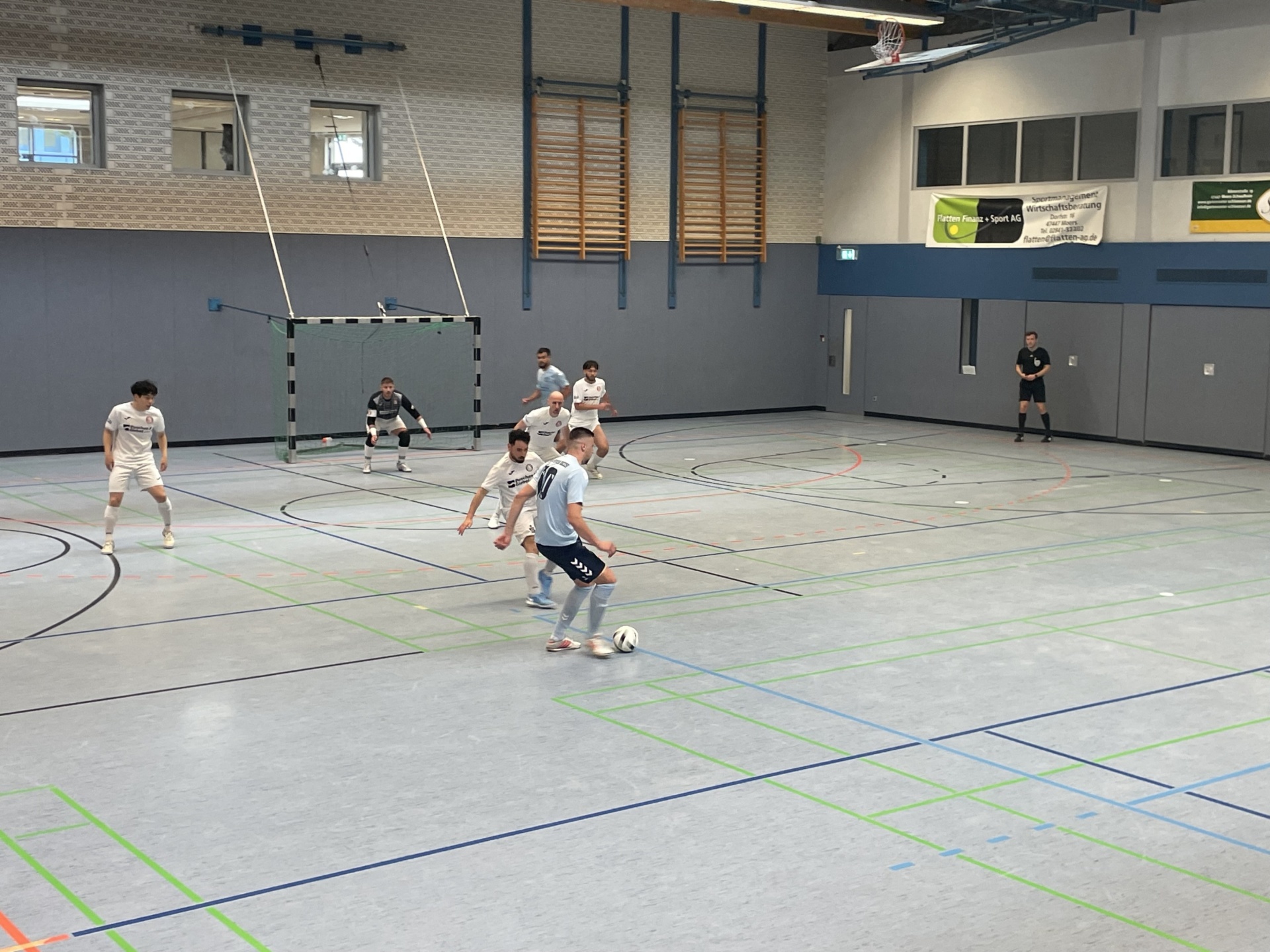 [{"clang_id":1,"value":"Handballspiel in Sporthalle. Spieler in hellen Trikots mit Ball, Gegner in dunkler Kleidung. Tore und Zuschauer im Hintergrund."}]