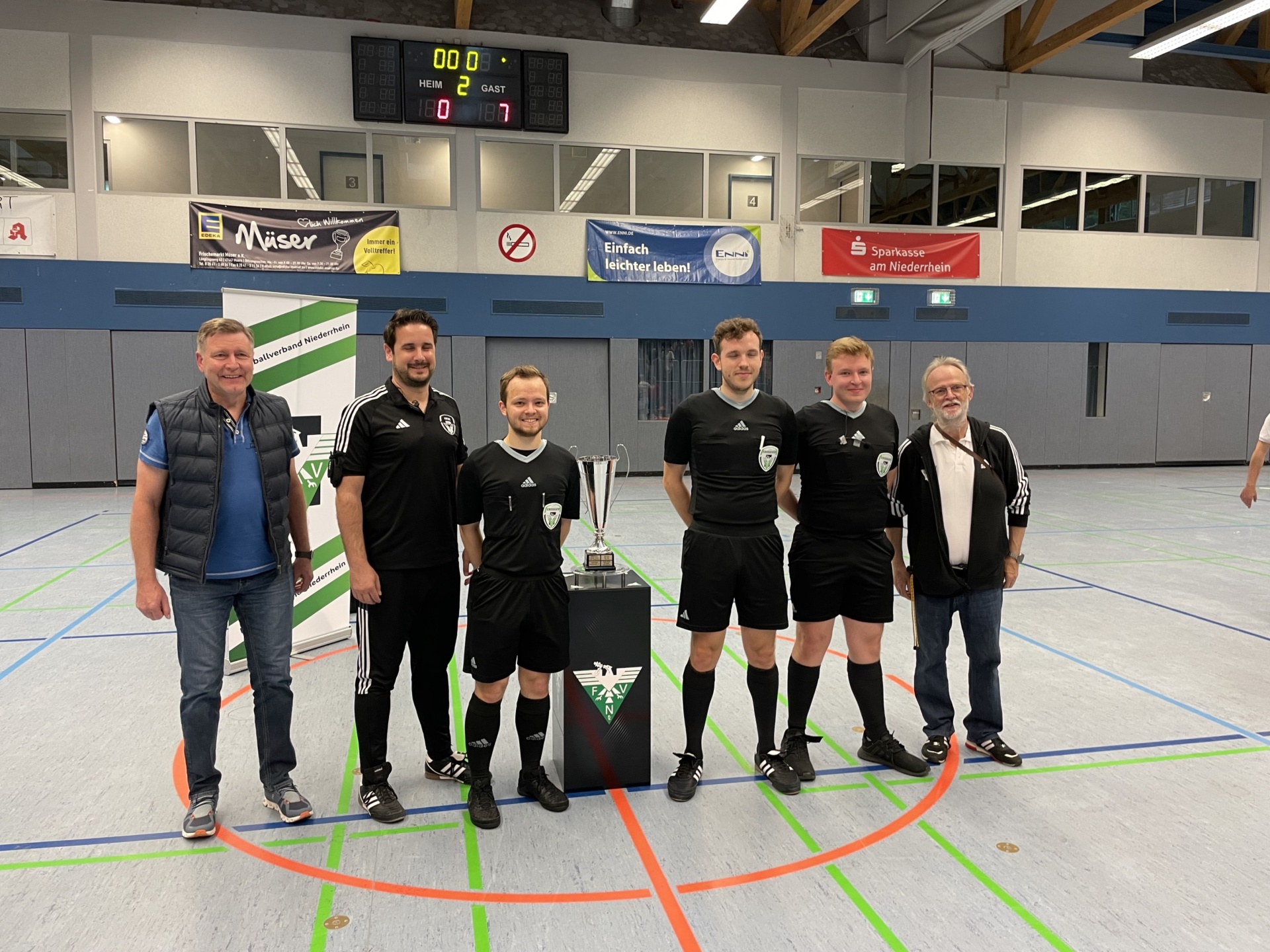 [{"clang_id":1,"value":"Gruppe von sechs Männern in Sportkleidung vor einer Sporthalle. Im Hintergrund eine Anzeigetafel und Werbebanner. Ein Pokal steht zwischen ihnen."}]