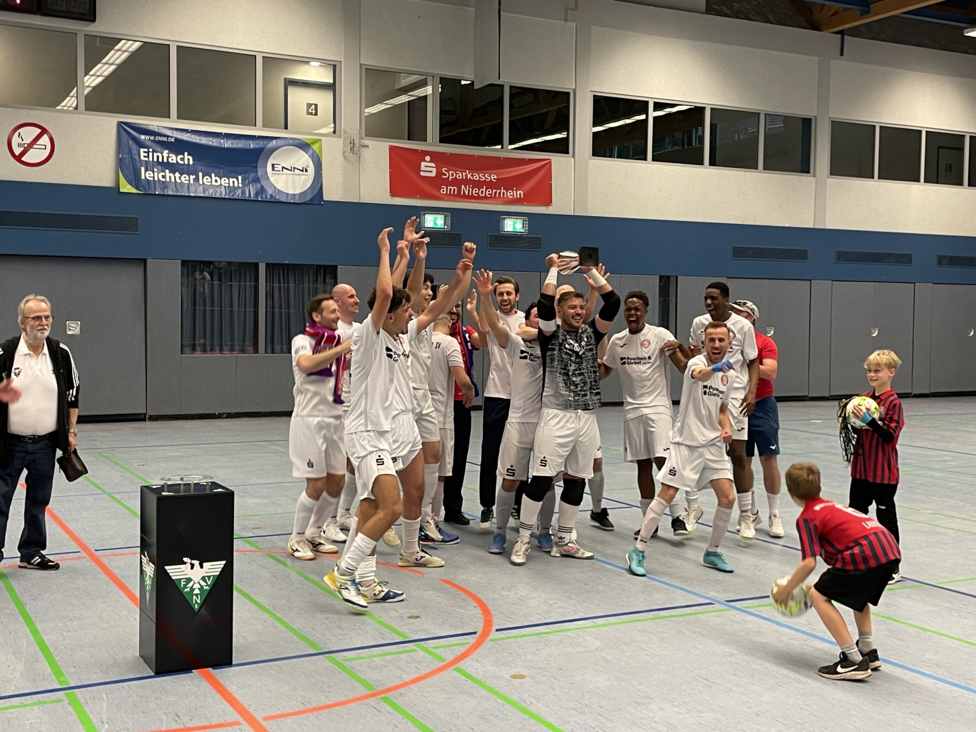 [{"clang_id":1,"value":"Handballteam in weißen Trikots jubelt mit verschränkten Armen. Im Hintergrund Zuschauer und Werbebanner in einer Sporthalle. Ein Kind wirft einen Ball."}]