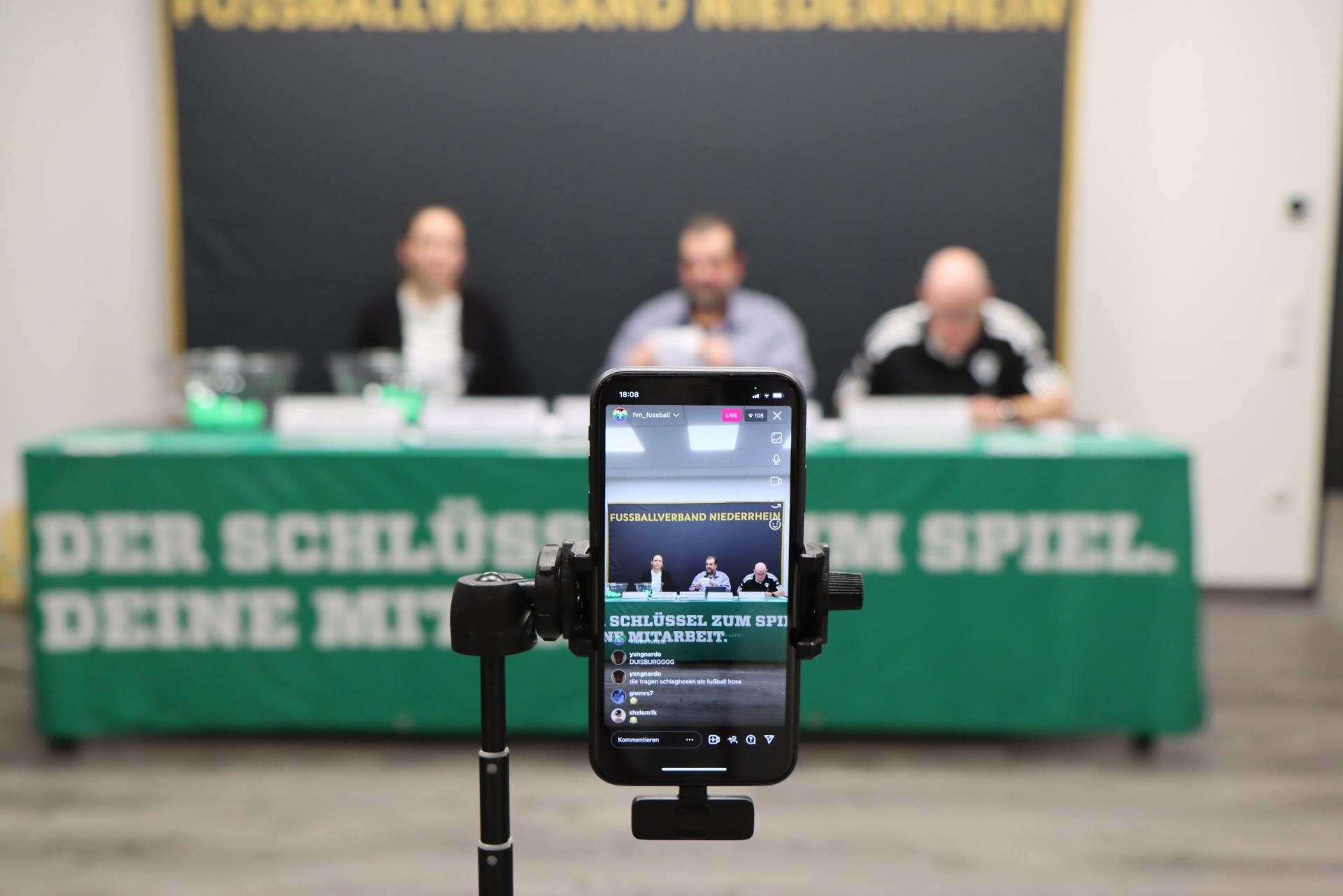 [{"clang_id":1,"value":"Konferenzraum mit Sitzendem Publikum. Im Vordergrund Smartphone auf Stativ, das einen Fußballverband-Bildschirm anzeigt. Banner mit Text im Hintergrund."}]