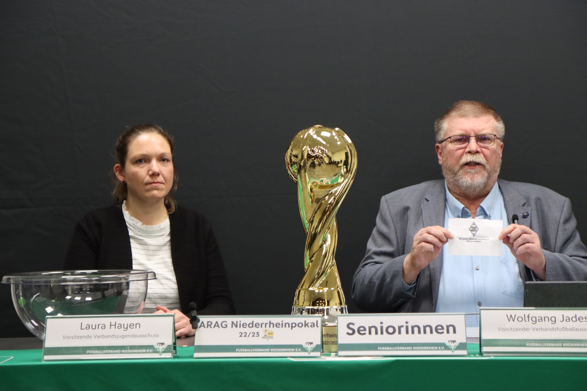 [{"clang_id":1,"value":"Drei Personen sitzen an einem Tisch. Links eine Frau, in der Mitte eine goldene Trophäe, rechts ein Mann. Vor ihnen Namensschilder. Hintergrund ist eine dunkle Wand."}]