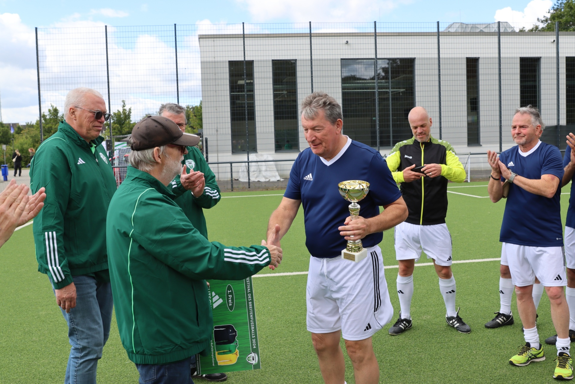 [{"clang_id":1,"value":"Eine Gruppe Männer auf einem Fußballfeld. Ein Mann übergibt einen Pokal an einen anderen, während die anderen applaudieren. Alle tragen Sportkleidung, meist in Grün und Weiß. Im Hintergrund ein modernes Gebäude."}]