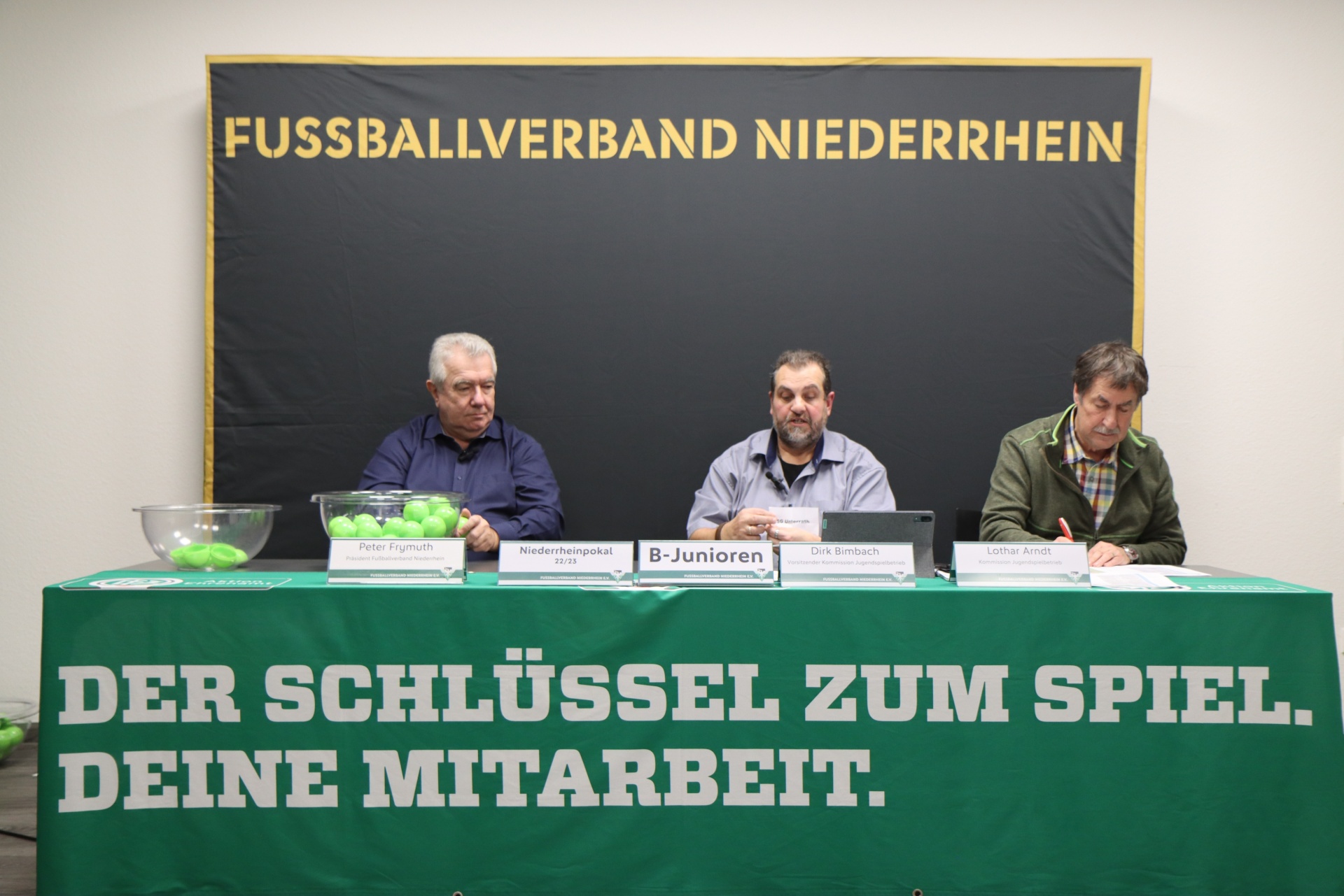 [{"clang_id":1,"value":"Konferenz-Szene: Drei Männer sitzen an einem Tisch mit Namensschildern vor einem Banner des \"Fussballverband Niederrhein\". Im Hintergrund eine grüne Wand."}]