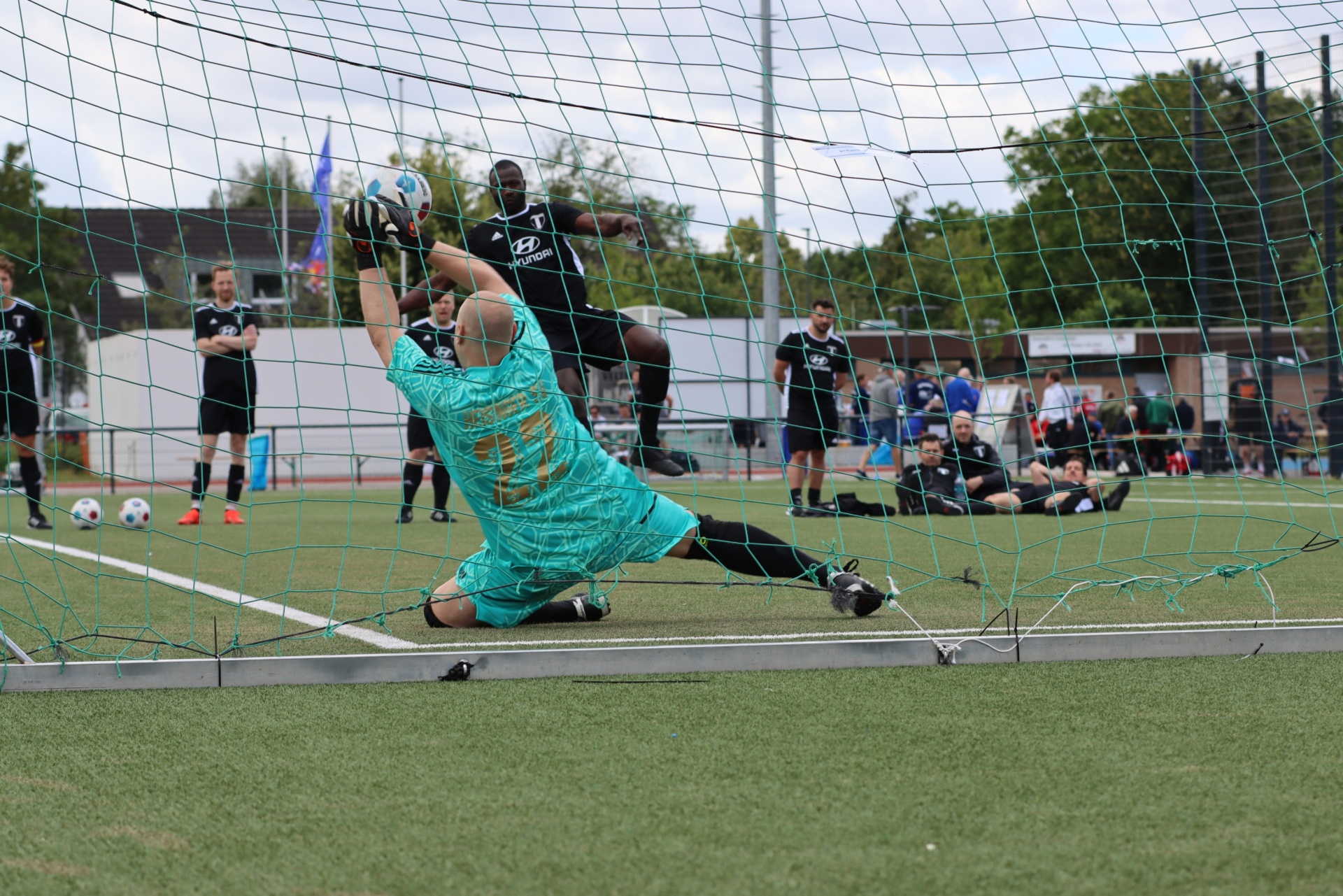 [{"clang_id":1,"value":"Fußballtor mit gerissenerem Netz. Torwart in Türkis/Schwarz springt zum Ball. Im Hintergrund stehen Fußballspieler auf grünem Rasen."}]