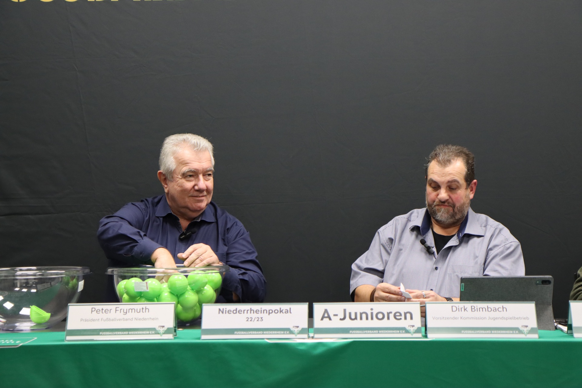 [{"clang_id":1,"value":"Zwei Männer sitzen an einem Tisch mit Namensschildern und einer Schale voller grüner Bälle. Im Hintergrund steht \"A-Junioren\" und \"Niederrheinpokal\"."}]