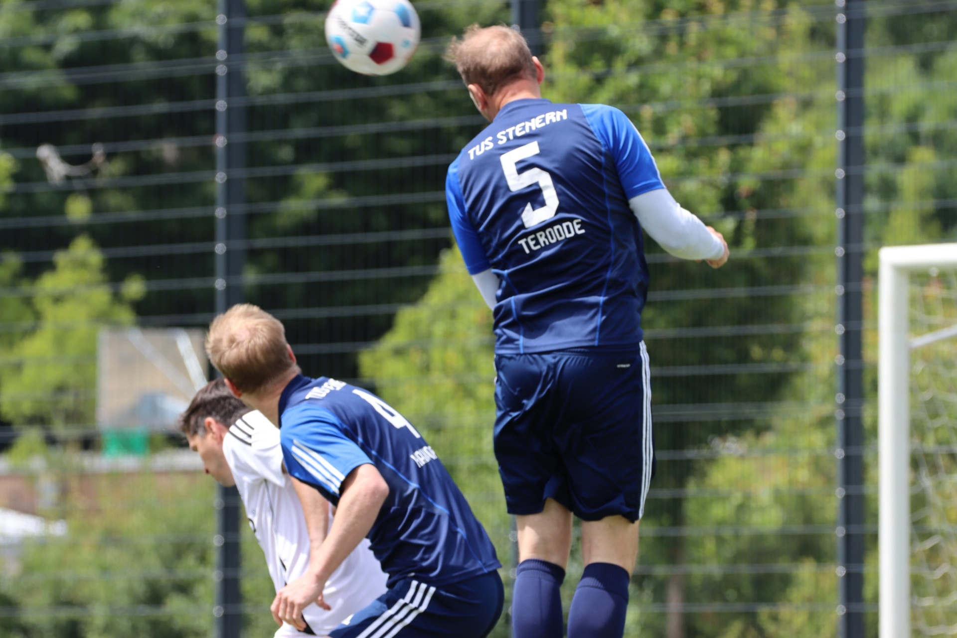 [{"clang_id":1,"value":"Fußballspieler mit Trikotnummer 5 springt zum Kopfball. Im Hintergrund weitere Spieler und ein Fußballtor. Grasplatz und Zaun im Hintergrund."}]