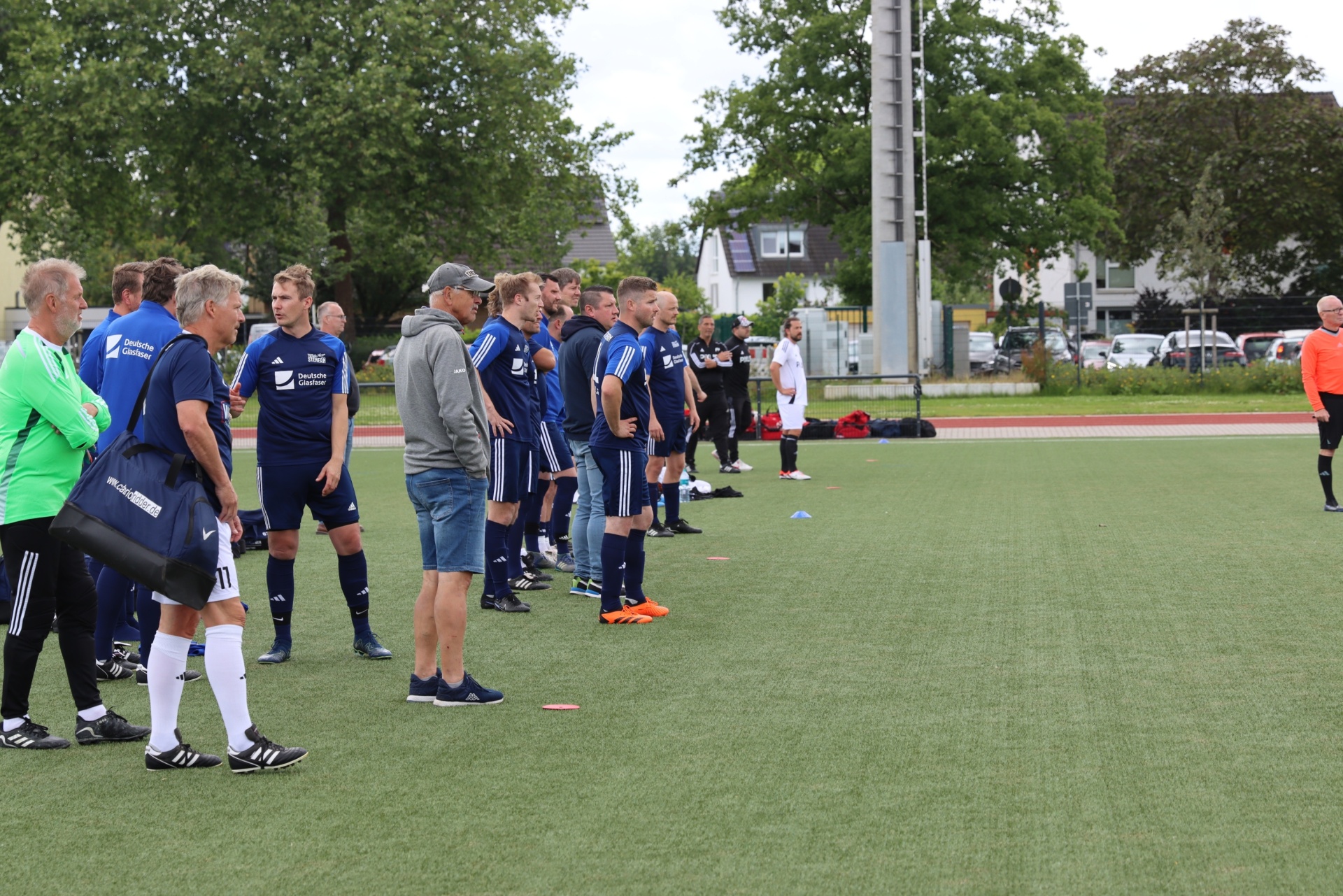 [{"clang_id":1,"value":"Fußballteam in blauen Trikots und Shorts steht auf grünem Rasen. Trainer und Betreuer stehen am Spielfeldrand, einige mit Sporttaschen. Gebäude im Hintergrund."}]