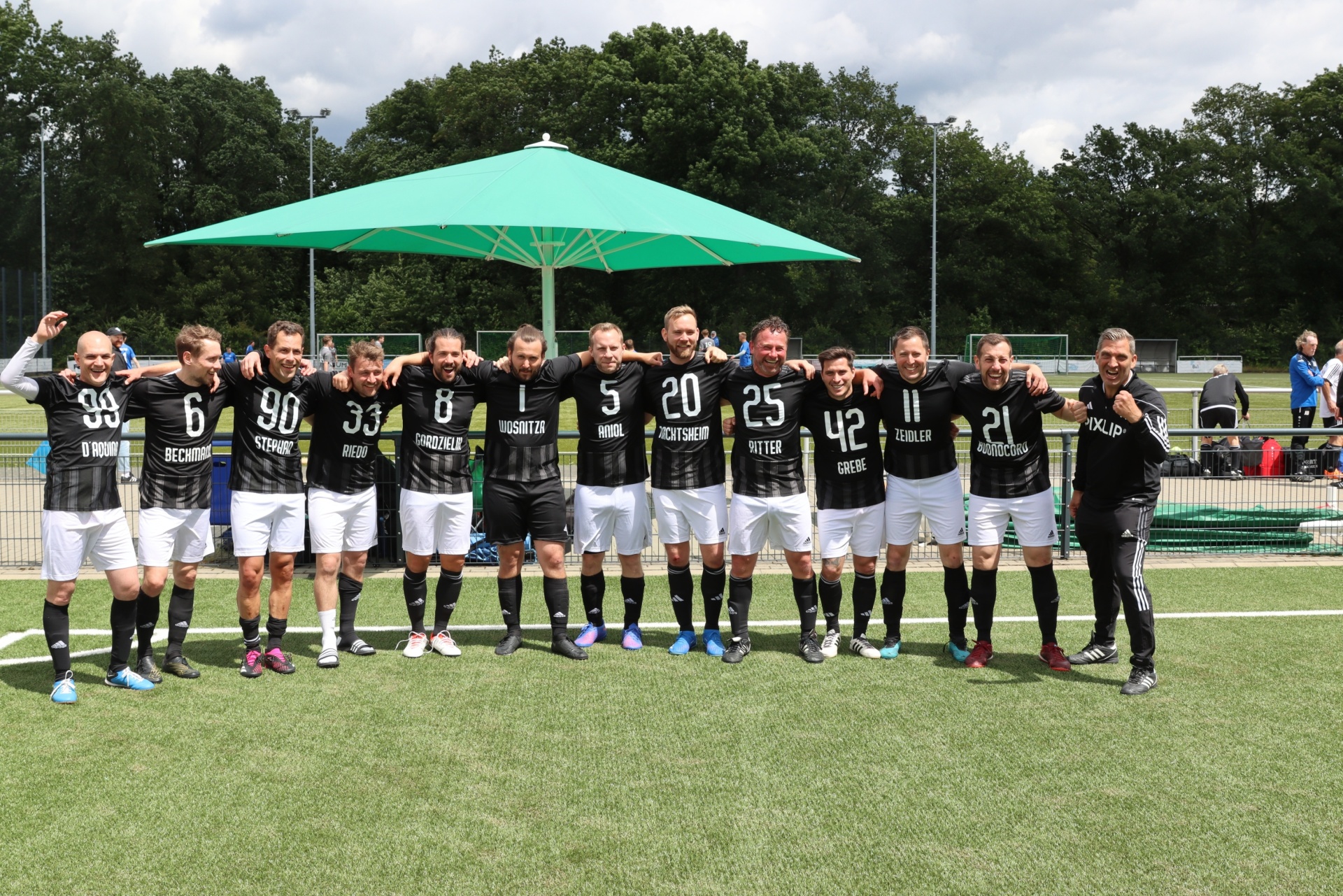 [{"clang_id":1,"value":"Fußballmannschaft in schwarzen Trikots und weißen Hosen steht auf einem grünen Rasenfeld vor einem grünen Zeltdach. Einige Personen stehen am Spielfeldrand."}]