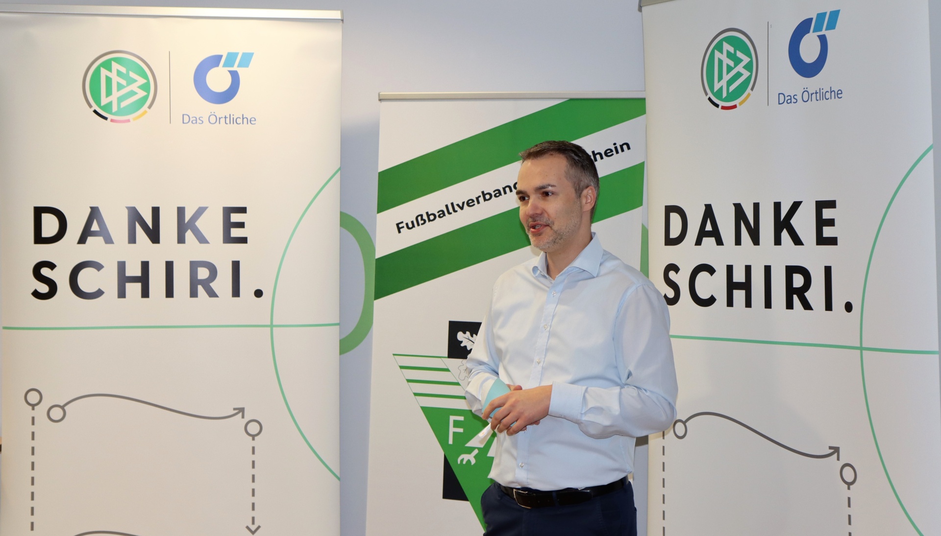 [{"clang_id":1,"value":"Mann mit Smartphone vor Bannern mit grünem Fussball-Design und Aufschrift \"Danke Schiri\". Im Hintergrund weiteres Banner mit Fussballverband-Schriftzug."}]