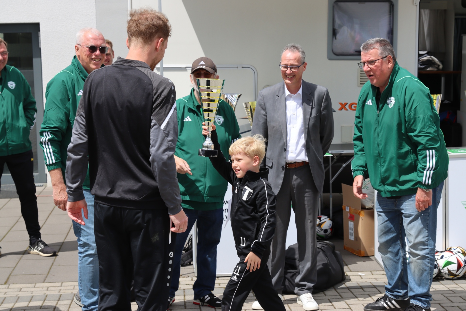 [{"clang_id":1,"value":"Fußballspieler mit Pokal übergibt ihn einem jungen Fan. Im Hintergrund stehen weitere Personen in Trainingsanzügen und ein Mann im Anzug."}]