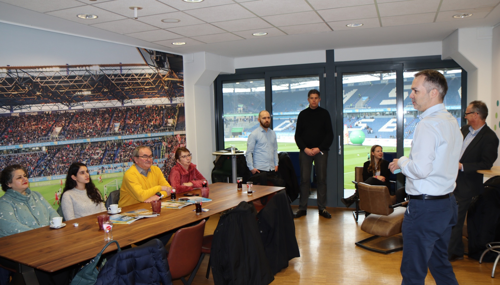 [{"clang_id":1,"value":"Besprechungsraum mit Blick auf Fußballstadion. Eine Gruppe von etwa zehn Personen sitzt oder steht um einen großen Konferenztisch. Ein Mann spricht und gestikuliert."}]