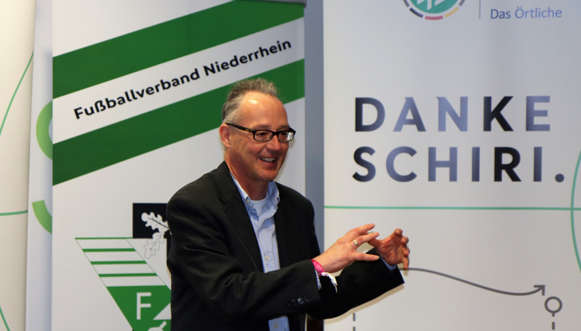 [{"clang_id":1,"value":"Mann mit Brille und schwarzem Jackett vor Hintergrund mit Text \"Fußballverband Niederrhein\" und \"DANKE SCHIRI\". Er gestikuliert mit beiden Händen."}]