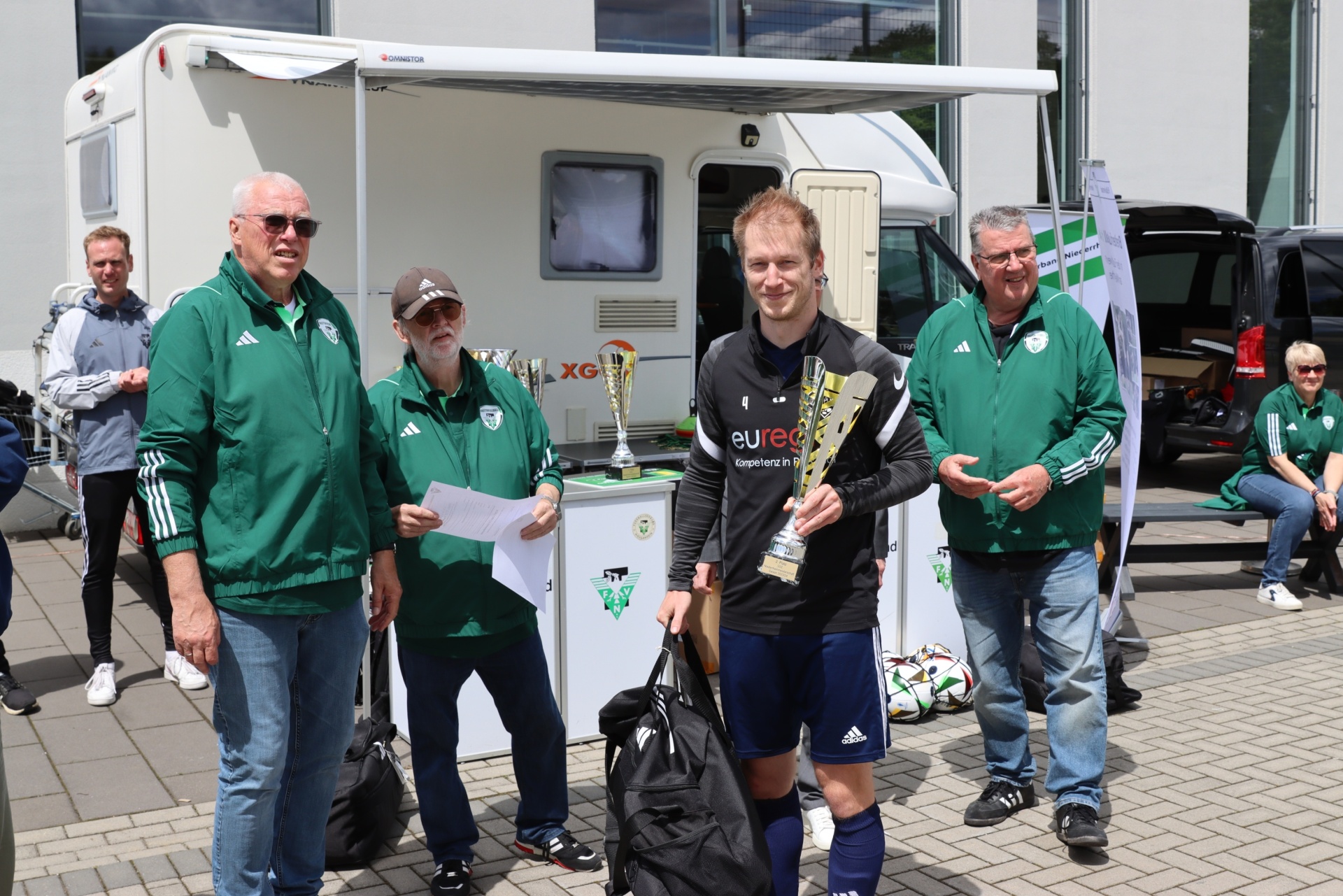 [{"clang_id":1,"value":"Fünf Männer stehen vor einem Wohnmobil. Ein junger Mann in Sportkleidung hält zwei Pokale. Die anderen tragen grüne Jacken und Jeans. Im Hintergrund weitere Personen und Fahrzeuge."}]