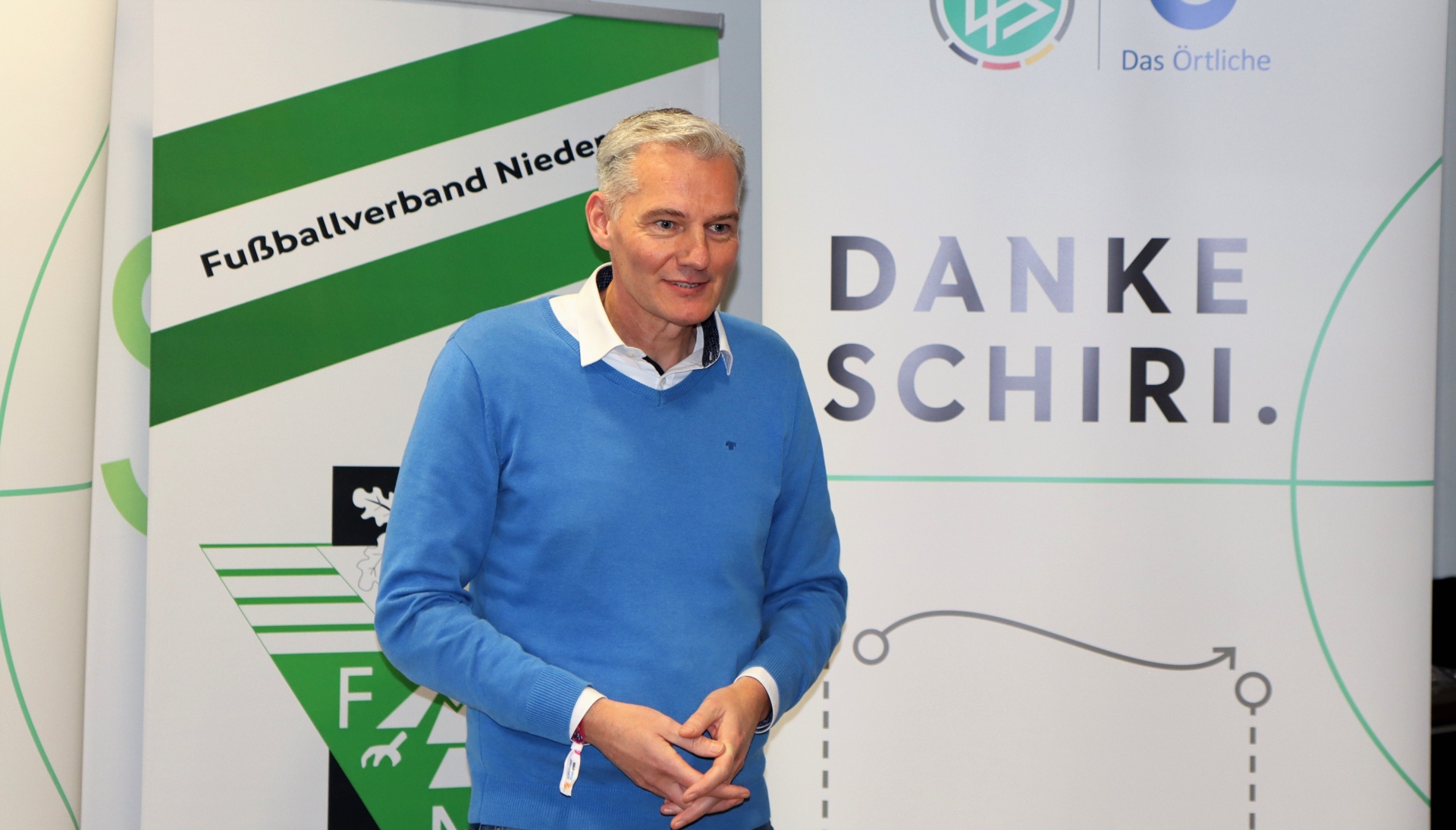 [{"clang_id":1,"value":"Mann in blauem Pullover vor Hintergrund mit Fussball-Logo und Schriftzug \"DANKE SCHIRI\". Fussballfeld-Illustration im Hintergrund."}]