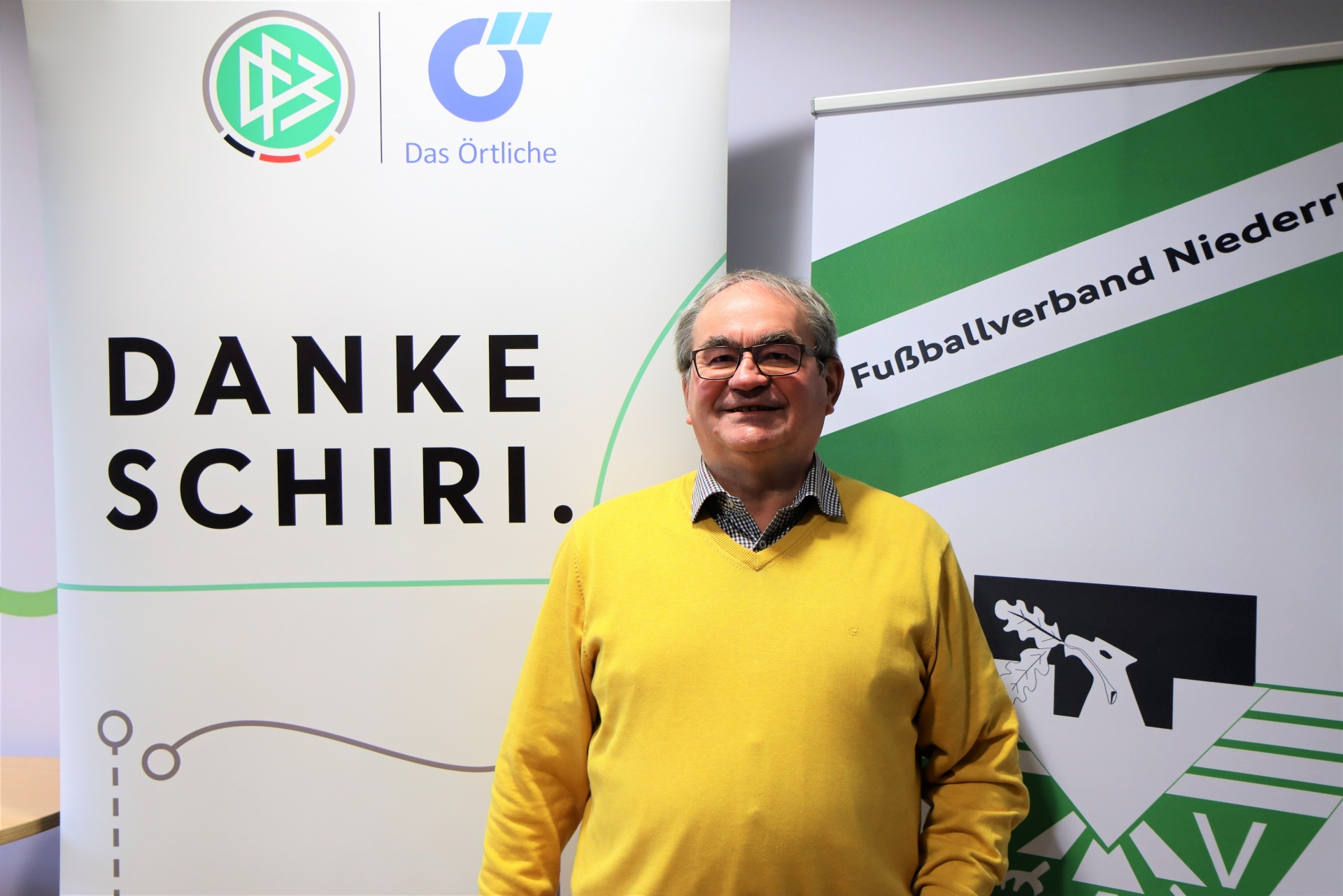[{"clang_id":1,"value":"Mann mit Brille und gelbem Pullover vor Werbebanner mit Schriftzug \"DANKE SCHIRI.\" und Logo des Fußballverbands Niederrhein."}]