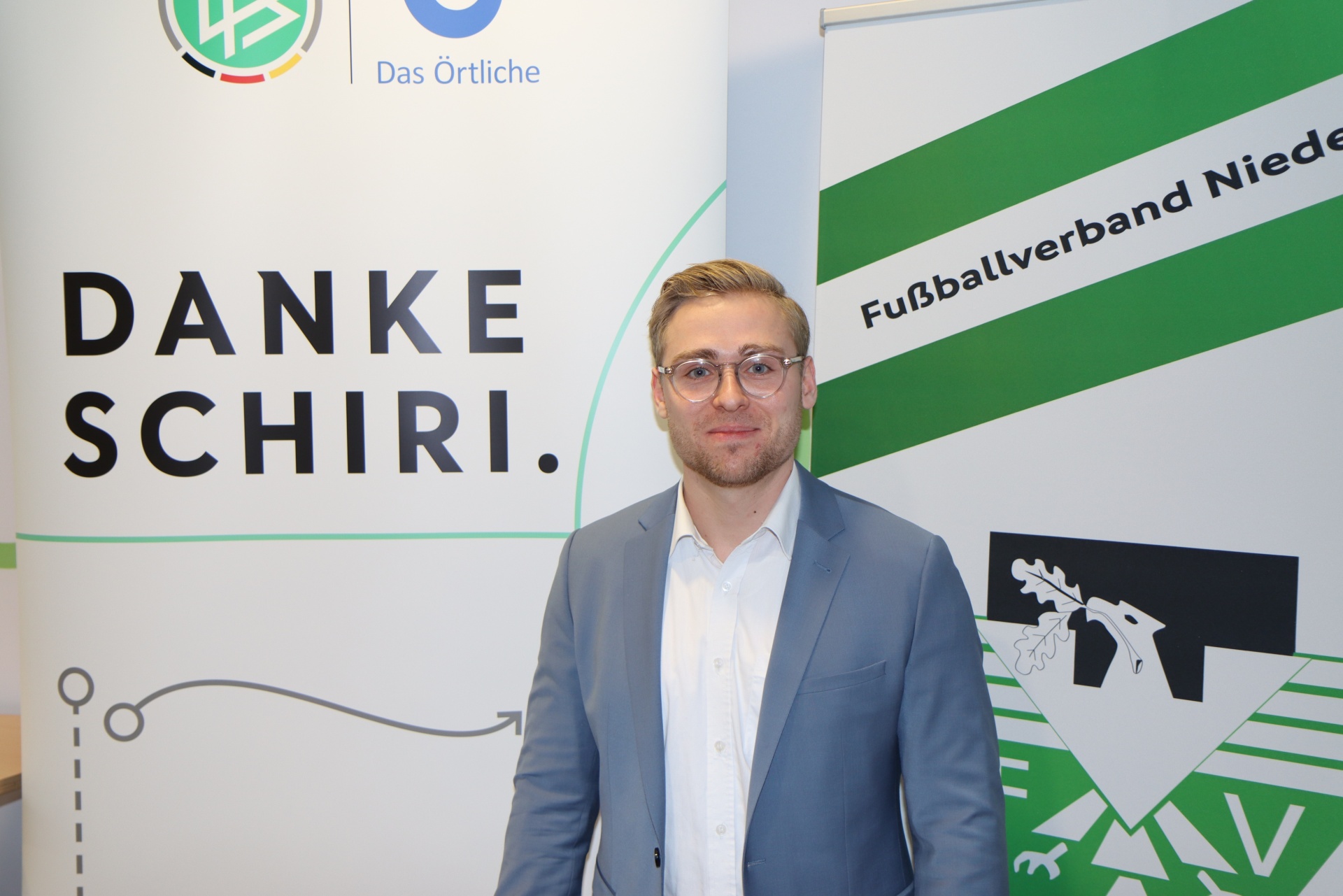 [{"clang_id":1,"value":"Mann mit blonden Haaren und Brille in grauem Anzug vor Hintergrund mit Schriftzug \"DANKE SCHIRI.\" und Fußballverbandslogo."}]