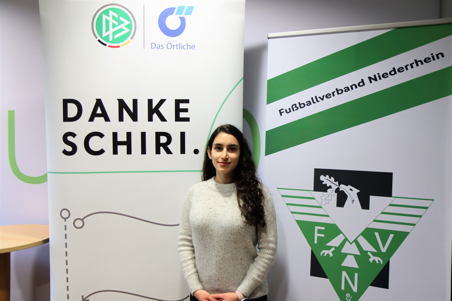 [{"clang_id":1,"value":"Frau mit lockigem Haar vor Werbebanner mit Schriftzügen \"Danke Schiri.\" und \"Fußballverband Niederrhein.\" sowie Logos."}]