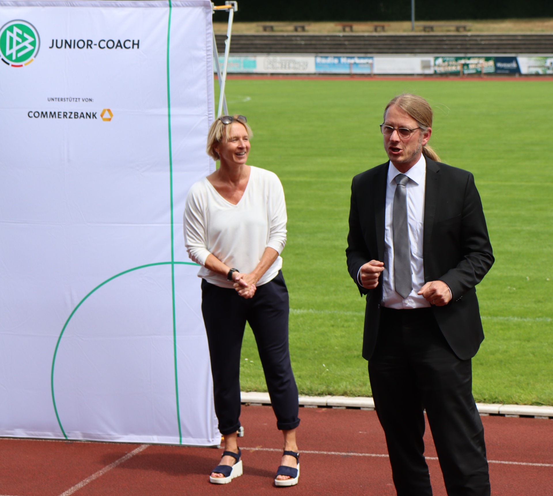 [{"clang_id":1,"value":"Zwei Personen vor einem Werbebanner mit dem Schriftzug \"JUNIOR-COACH\" und dem Logo der Commerzbank auf einem Sportplatz. Eine Frau in legerer Kleidung lächelt, ein Mann im Anzug gestikuliert."}]