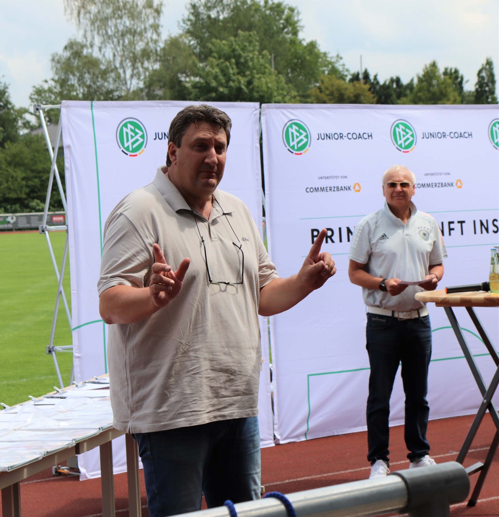 [{"clang_id":1,"value":"Zwei Männer stehen vor Bannern mit dem Schriftzug \"Junior Coach\". Der Mann links gestikuliert, der rechte steht entspannt da. Im Hintergrund ein Fußballfeld."}]