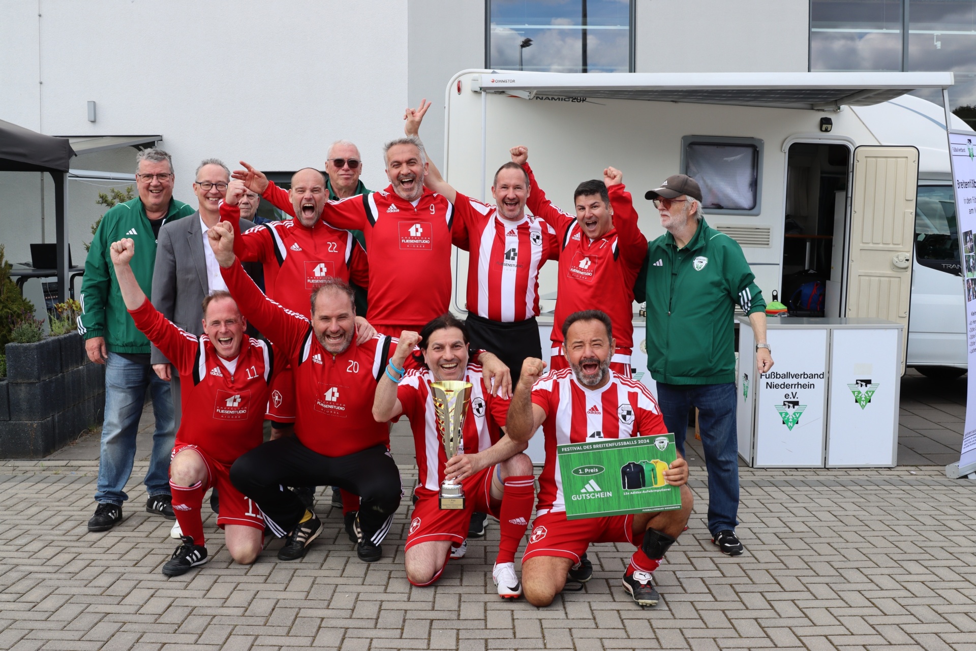 [{"clang_id":1,"value":"Fußballmannschaft in roten und weißen Trikots feiert Sieg mit Pokal. Im Hintergrund ein Wohnmobil und Personen in Freizeitkleidung."}]