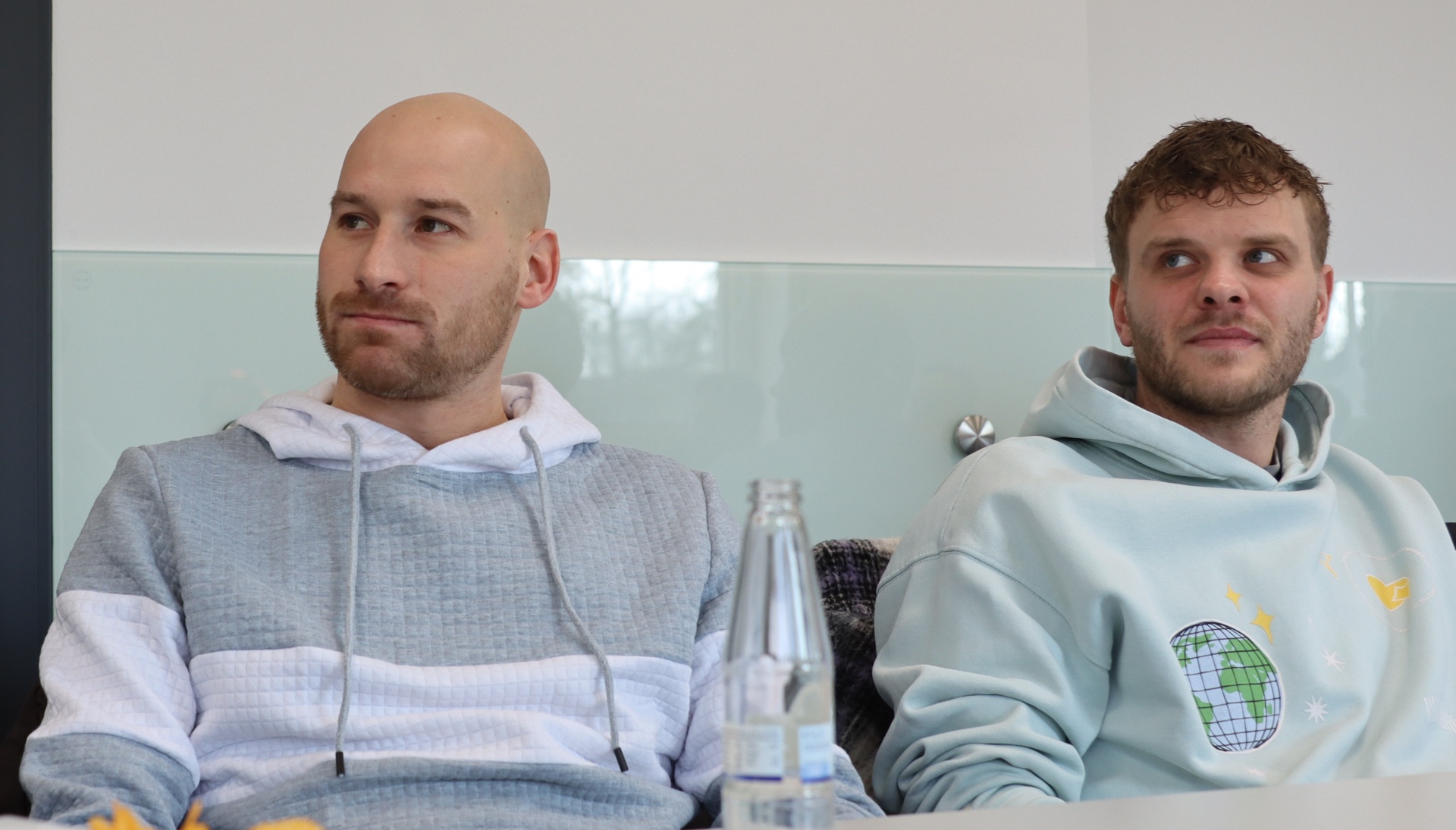 [{"clang_id":1,"value":"Zwei junge Männer sitzen nebeneinander vor einer hellen Wand. Links ein Mann mit Glatze und blauem Hoodie, rechts ein Mann mit dunklem Haar und hellblauem Hoodie mit Weltkugel-Motiv. Zwischen ihnen eine Wasserflasche."}]