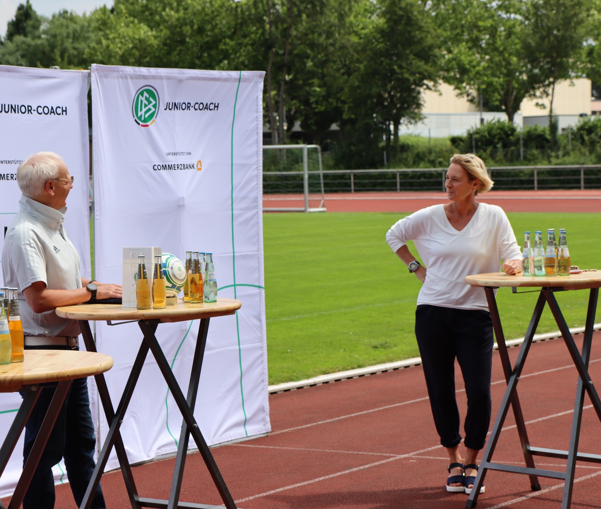 [{"clang_id":1,"value":"Zwei Personen stehen an Stehtischen vor einem Banner mit dem Logo \"JUNIOR-COACH\" und dem Sponsor \"Commerzbank\" neben einem Sportfeld. Getränke sind auf den Tischen platziert."}]