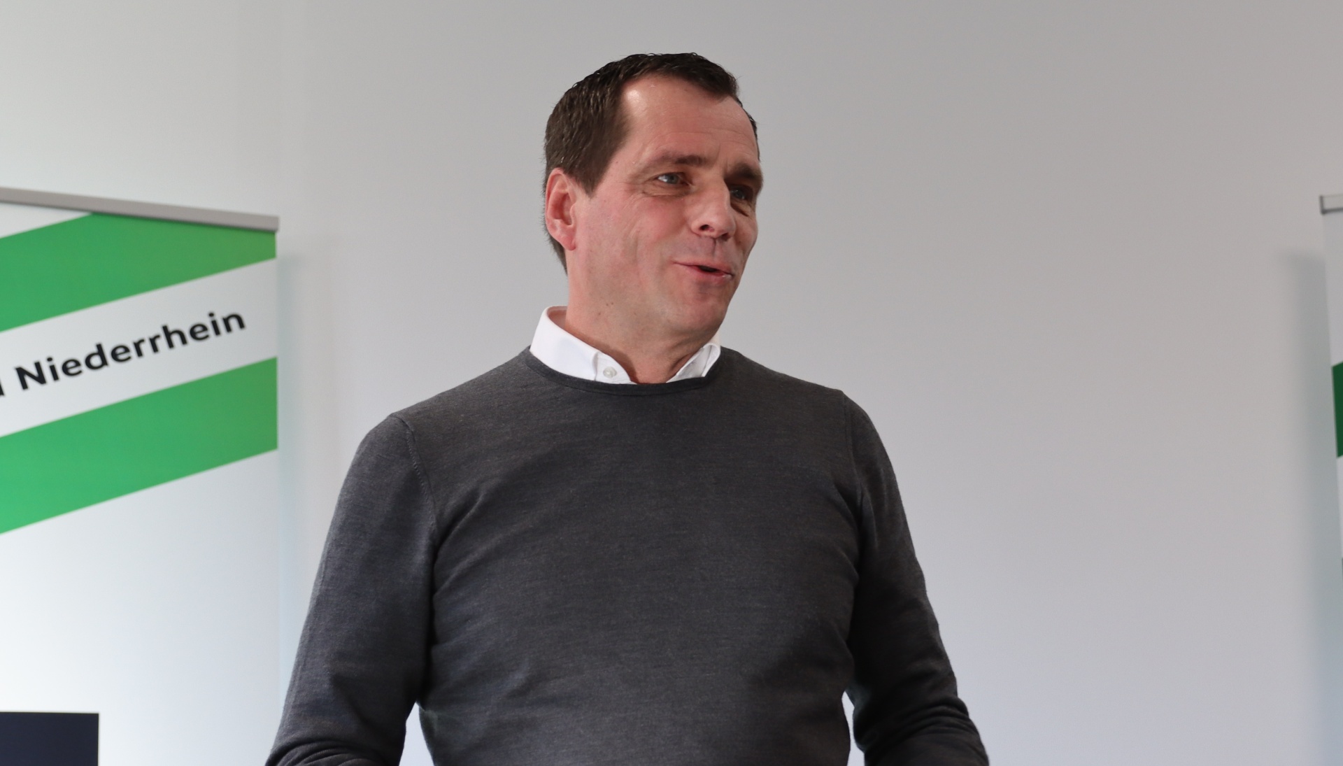 [{"clang_id":1,"value":"Mann mit braunen Haaren, grauem Pullover und weißem Hemd vor grünem Hintergrund mit Schrift \"Niederrhein\"."}]