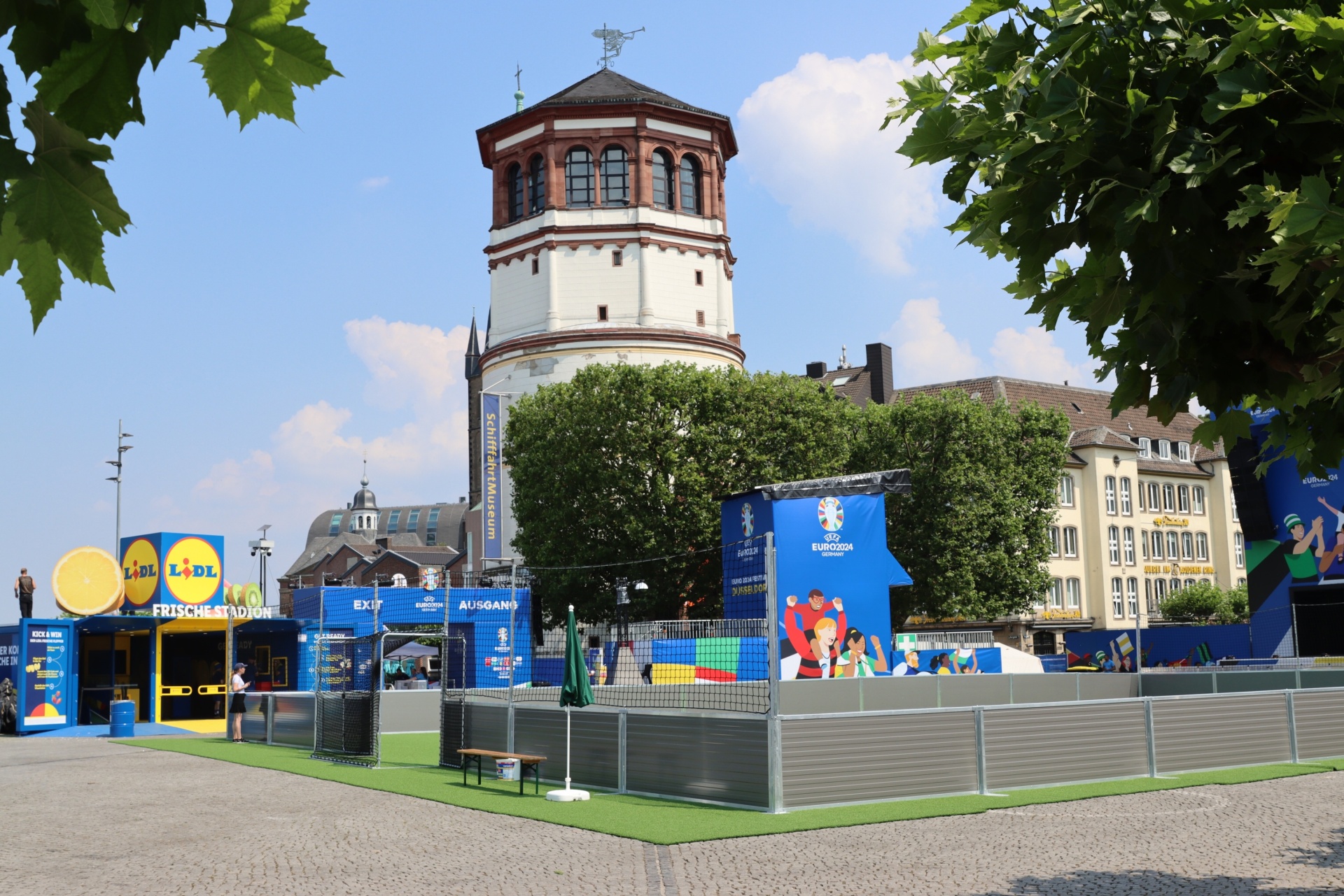 [{"clang_id":1,"value":"Stadtplatz mit mittelalterlichem Wehrturm, umgeben von Gebäuden. Im Vordergrund Absperrungen und Werbeaufsteller für ein Sportereignis. Blätterbäume säumen den Rand."}]