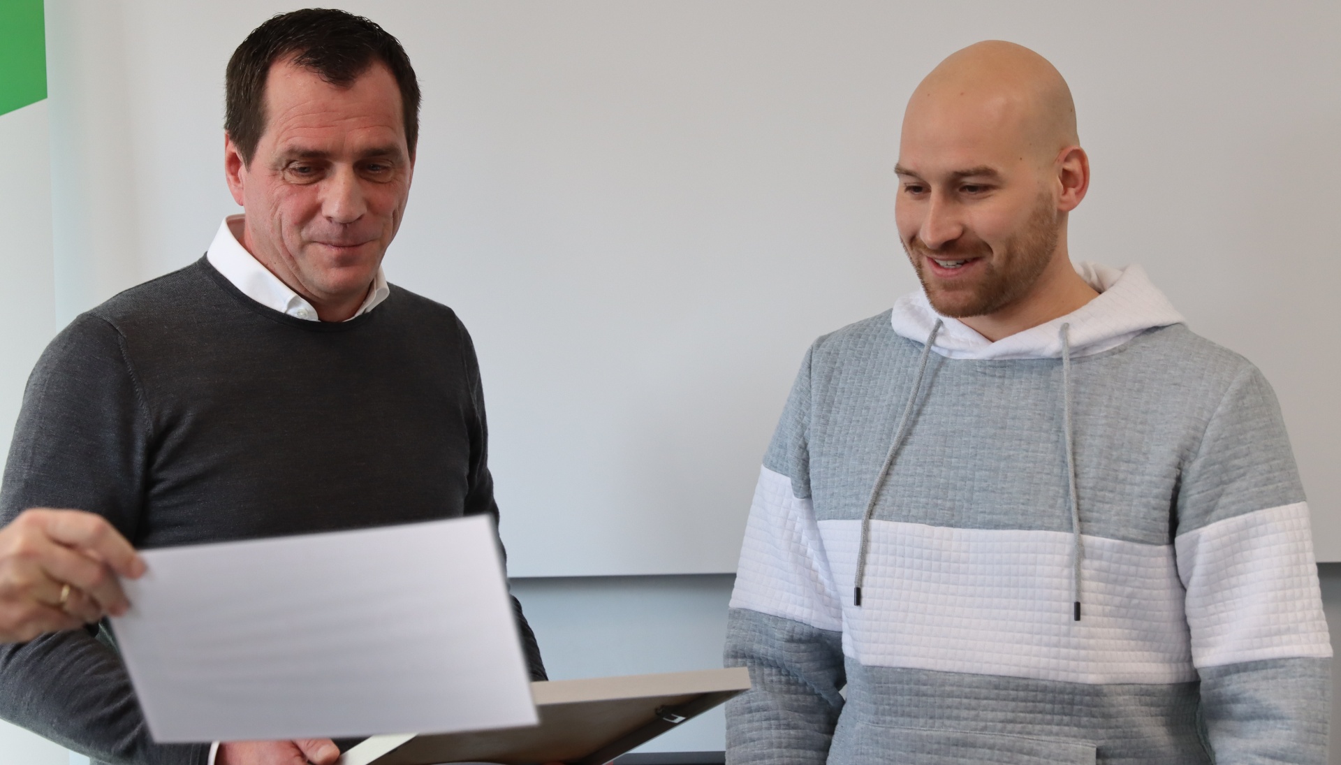 [{"clang_id":1,"value":"Zwei Männer mittleren Alters betrachten gemeinsam ein Dokument. Der linke trägt Pullover, der rechte eine Jacke mit Kapuze. Im Hintergrund eine weiße Wand."}]