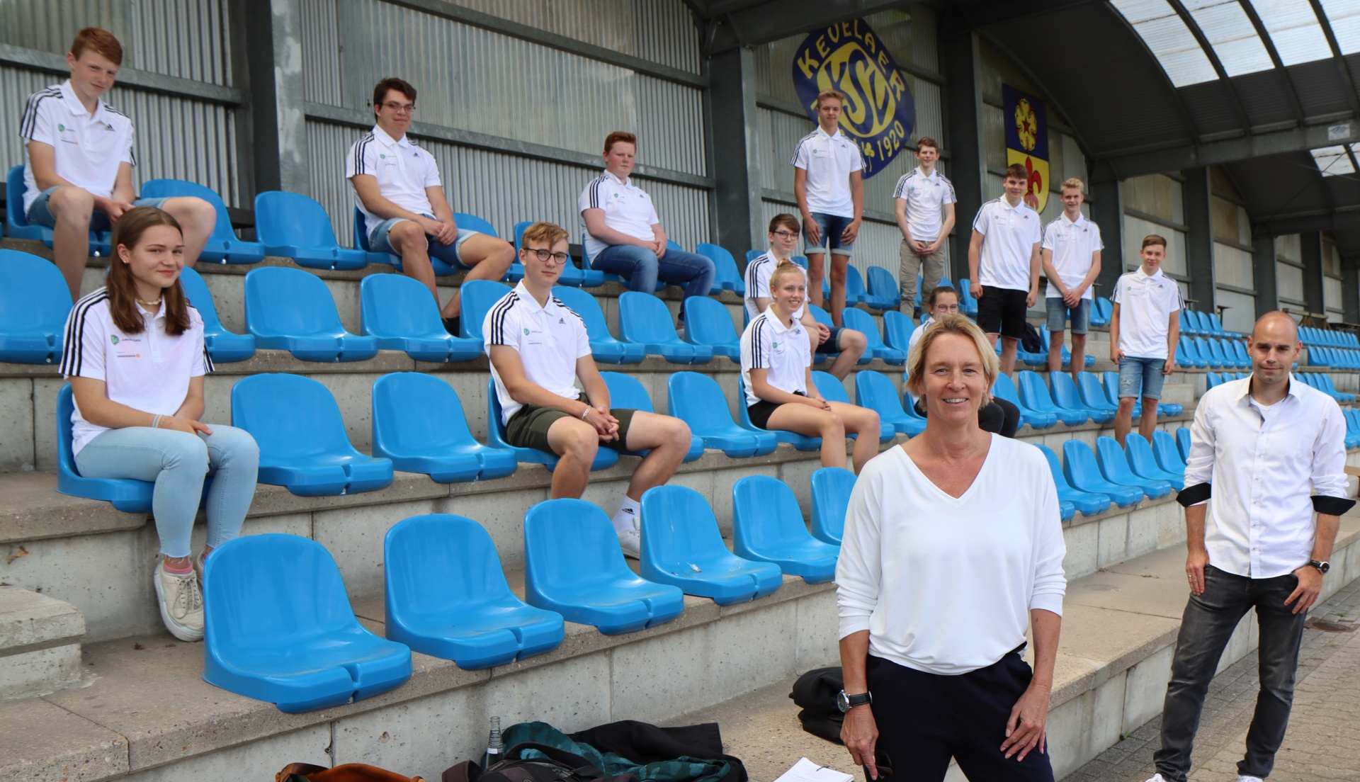 [{"clang_id":1,"value":"Gruppe junger Menschen und zwei Erwachsene sitzen auf blauen Stadiontribünen vor einem Fußballstadion. Alle tragen helle Oberteile."}]