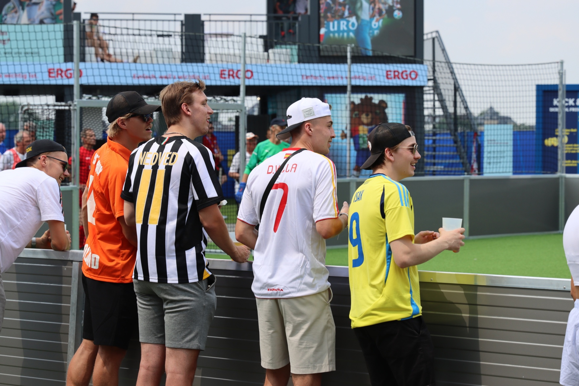 [{"clang_id":1,"value":"Fünf Männer stehen nebeneinander, tragen Fußballtrikots verschiedener Nationalitäten (Orange, Schwarz-Weiß, Rot, Blau-Gelb) und blicken in eine Richtung. Im Hintergrund eine Stadiontribüne und Werbebanner."}]