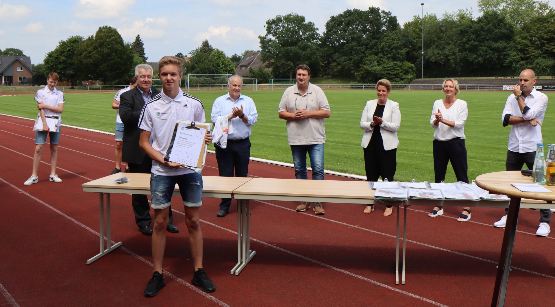 [{"clang_id":1,"value":"Junge erhält Urkunde auf Sportplatz. Im Hintergrund stehen Personen und Bäume. Rote Laufbahn."}]