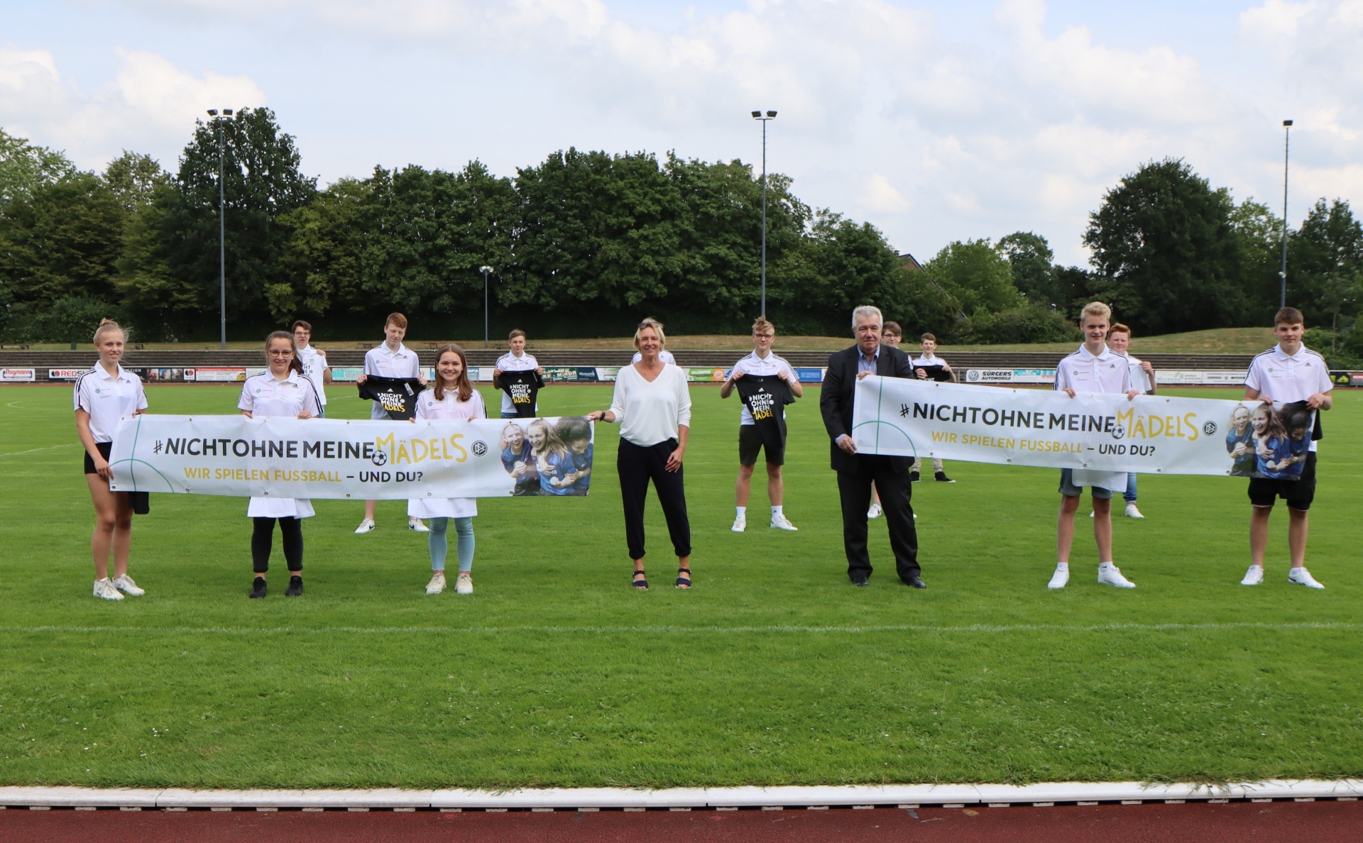 [{"clang_id":1,"value":"Fußballteam und zwei Erwachsene stehen auf grünem Rasen mit Banner: \"#NichtOhneMeineMädels – Wir spielen Fußball – und du?\". Im Hintergrund ein Fußballstadion."}]