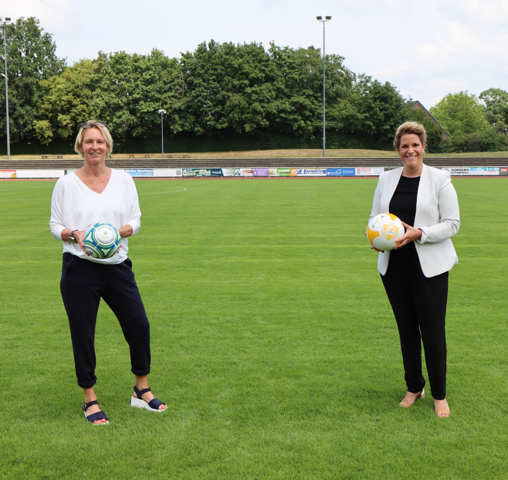 [{"clang_id":1,"value":"Zwei Frauen stehen auf einem Fußballfeld und halten jeweils einen Fußball. Im Hintergrund ein Stadion mit Zuschauerrängen und Bäumen."}]