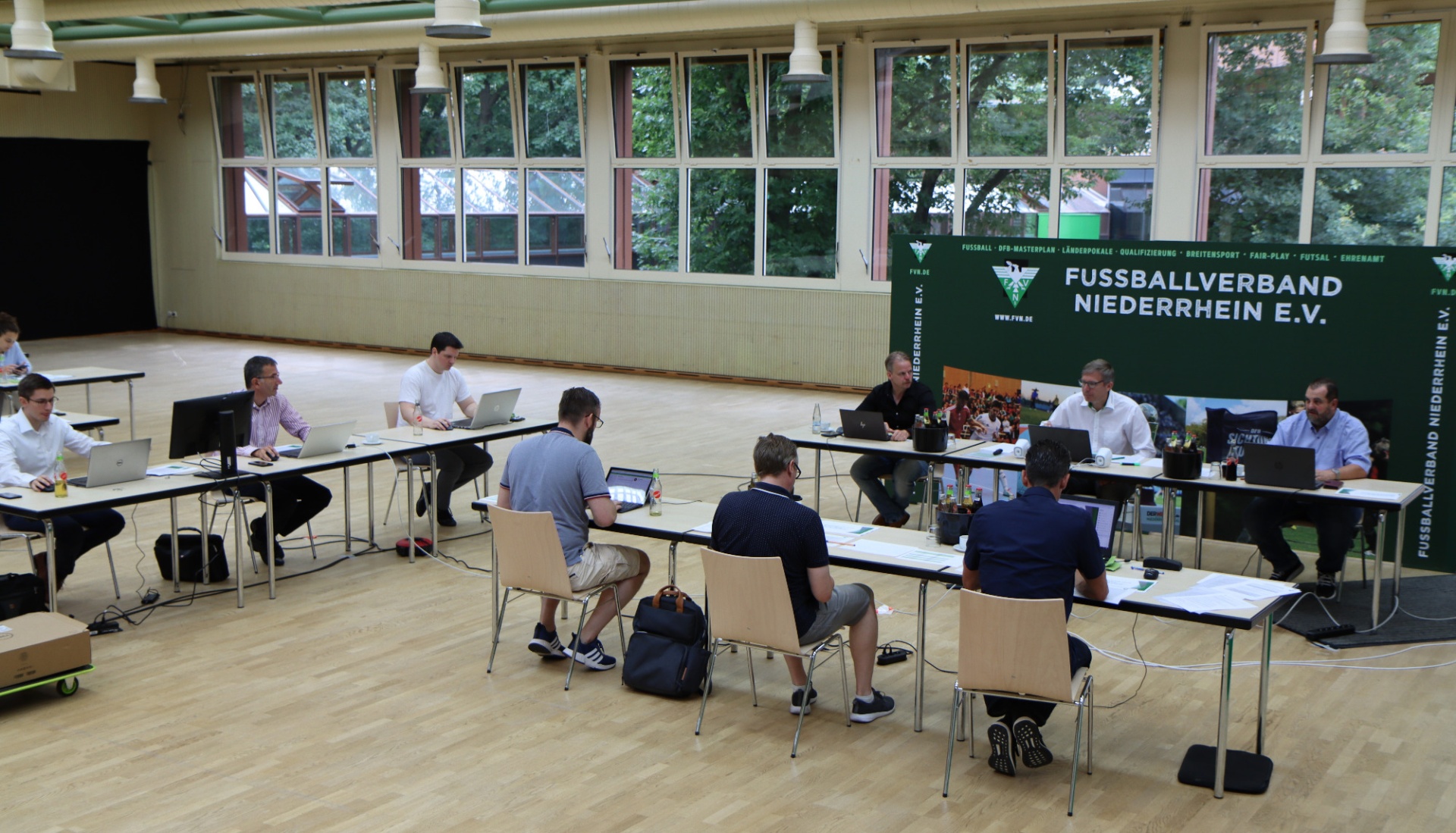 [{"clang_id":1,"value":"Versammlung in Sporthalle: Männer sitzen an Tischen, arbeiten mit Laptops und Papieren. Im Hintergrund Fenster mit Blick auf Bäume. Banner mit Fussballverband Niederrhein E.V."}]