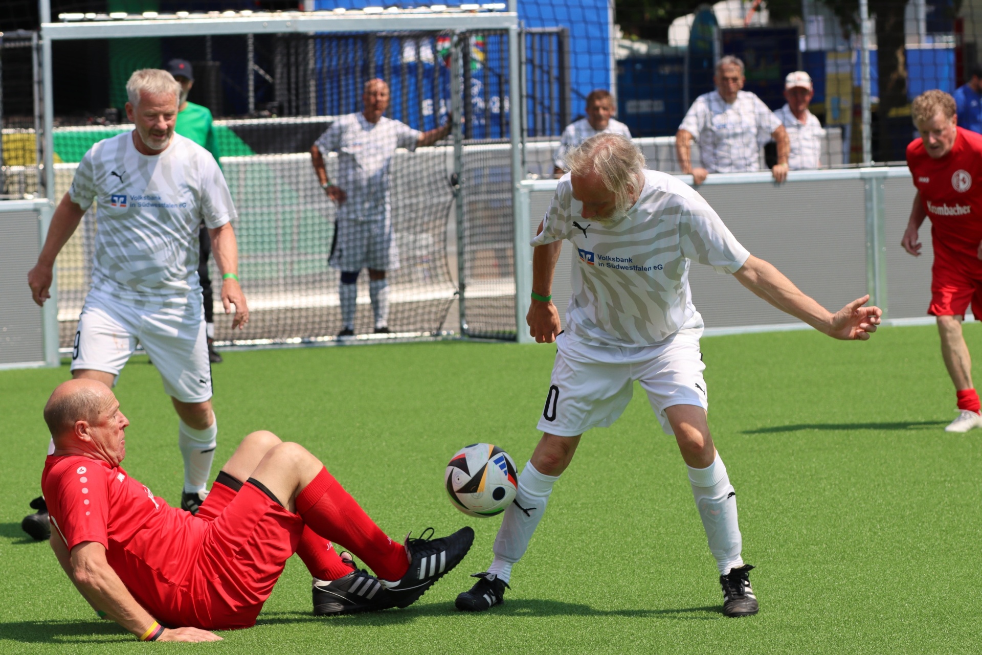 [{"clang_id":1,"value":"Fußballspiel: Spieler in weißen und roten Trikots auf grünem Kunstrasen. Ein Spieler tritt den Ball, ein anderer ist gefallen. Zuschauer im Hintergrund."}]
