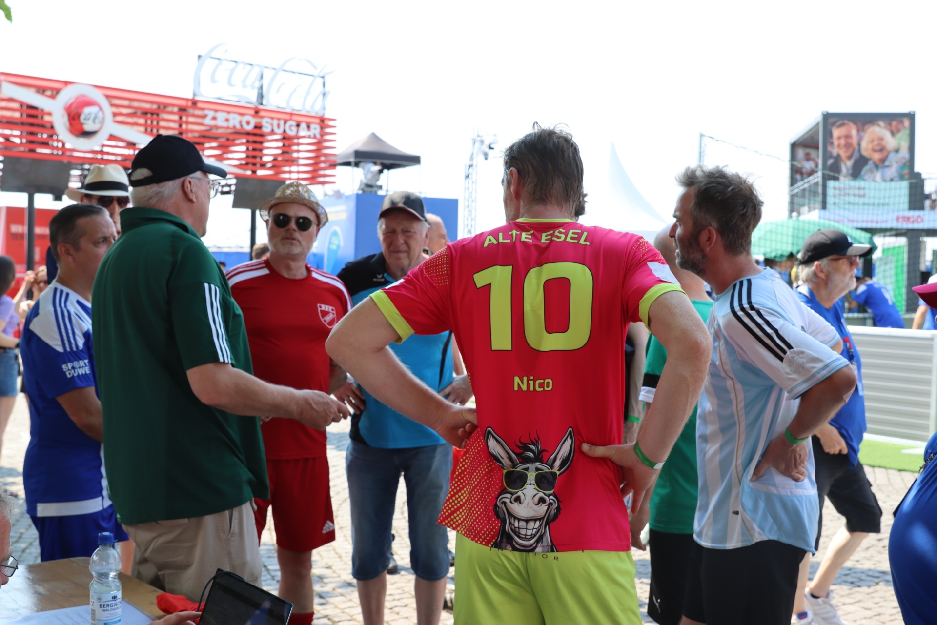 [{"clang_id":1,"value":"Fußballfans stehen in einer Menschenmenge. Ein Mann trägt ein rotes Trikot mit der Aufschrift \"ALTE ESEL\" und Nummer 10, sowie eine Hose mit Donkey-Motiv. Im Hintergrund Coca-Cola Werbung."}]