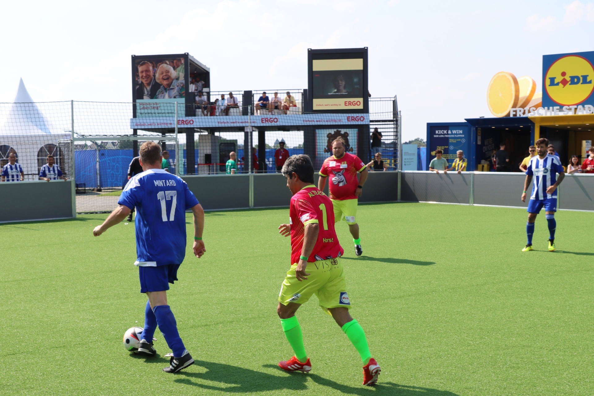 [{"clang_id":1,"value":"Fußballspiel im Freien: Zwei Spieler, einer blau, einer grün-gelb, kämpfen um den Ball. Im Hintergrund Zuschauer und Werbebanner."}]