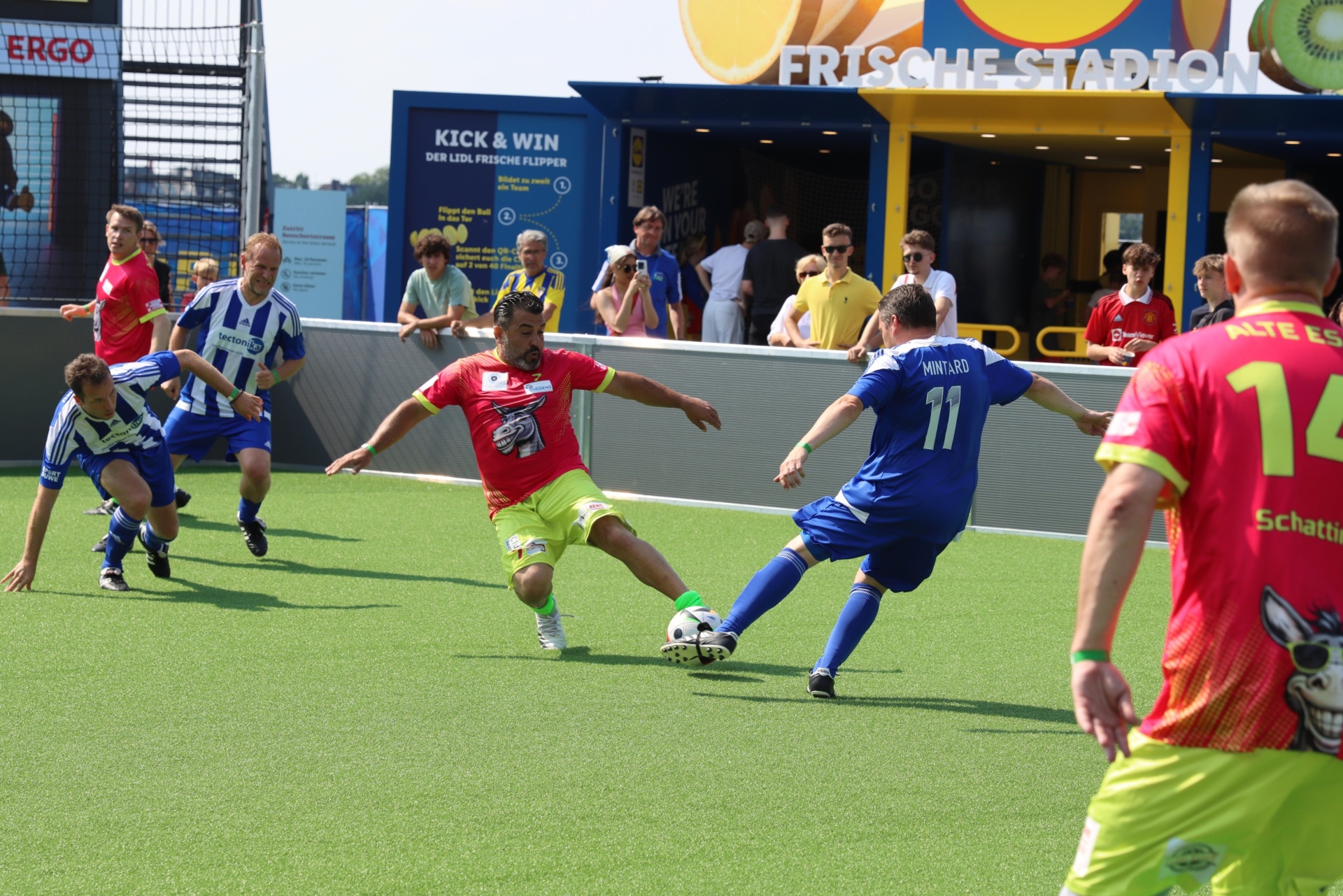 [{"clang_id":1,"value":"Fußballspiel auf grünem Kunstrasenfeld. Spieler in rot-gelben und blau-weißen Trikots kämpfen um den Ball. Im Hintergrund Zuschauer und Werbebanner."}]