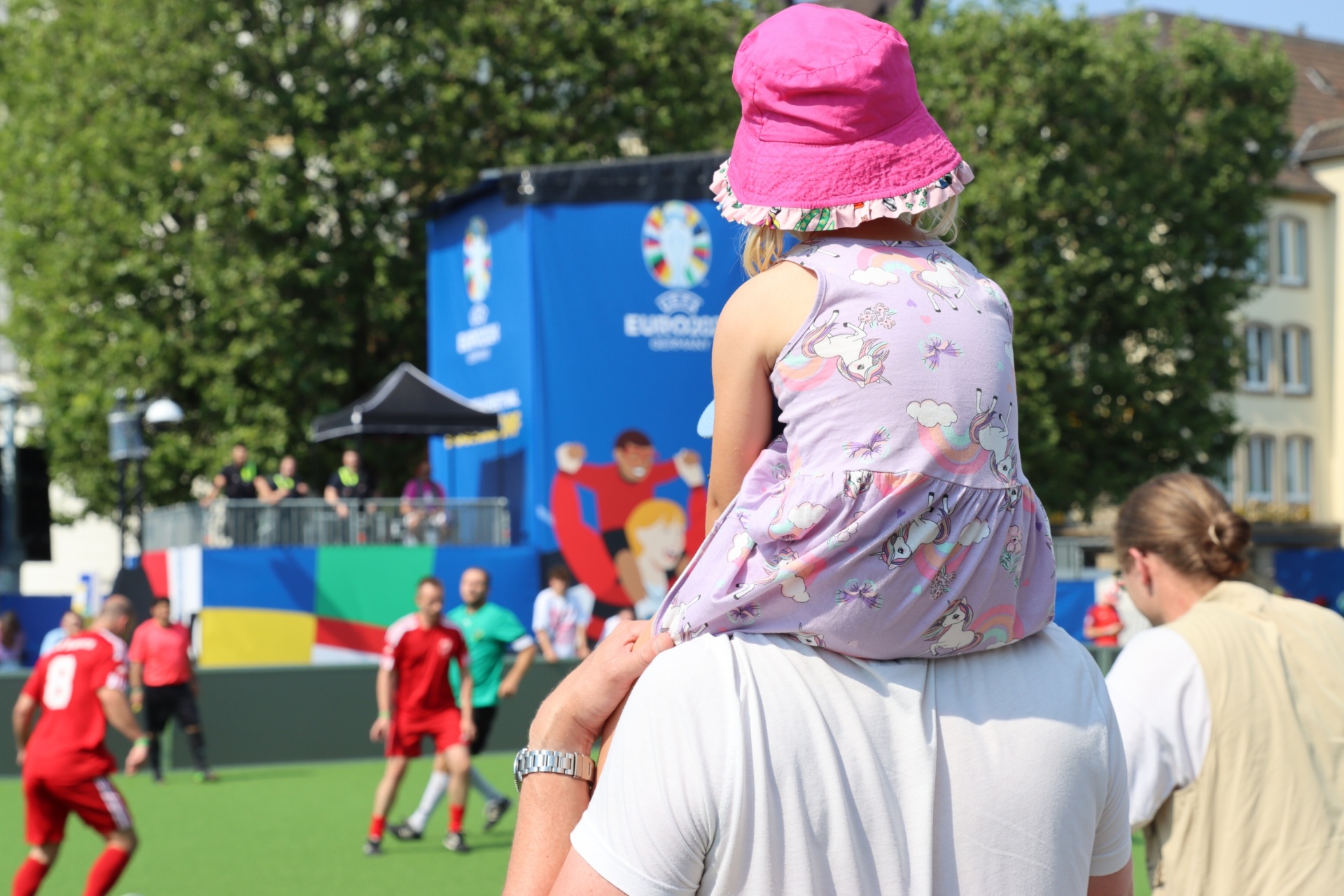 [{"clang_id":1,"value":"Ein Kind mit Einhorn-Kleid und pinkem Hut sitzt auf den Schultern einer Person. Im Hintergrund Fußballspieler und EURO 2024-Werbung."}]