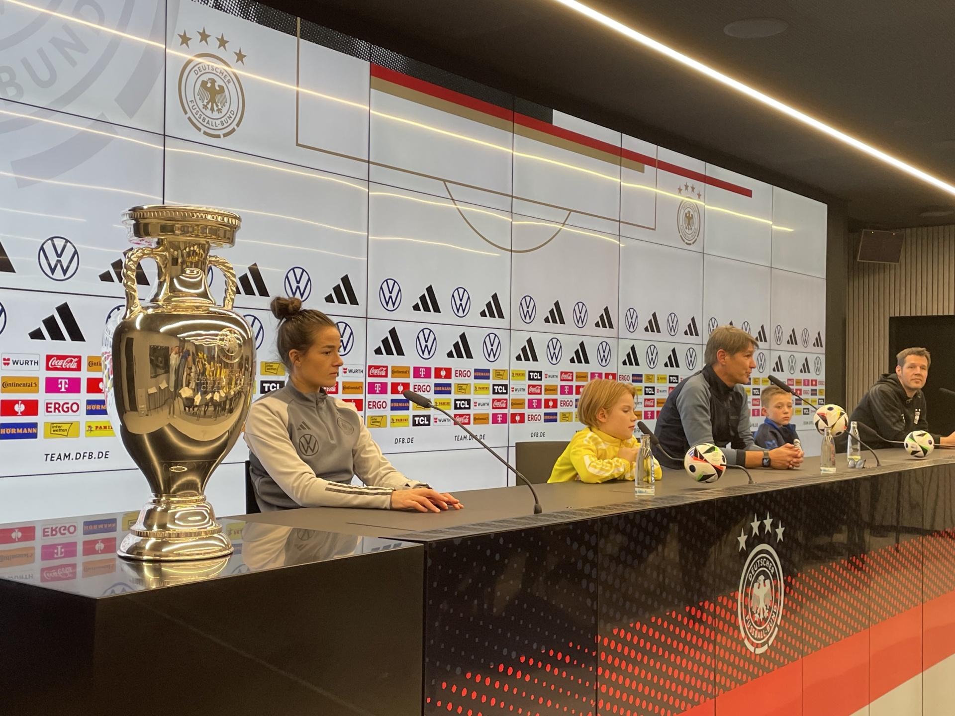 [{"clang_id":1,"value":"Frauenfußballteam sitzt an einem Tisch vor einem Hintergrund mit Fußballmotiven und dem DFB-Pokal. Eine Spielerin hält ein Handy."}]