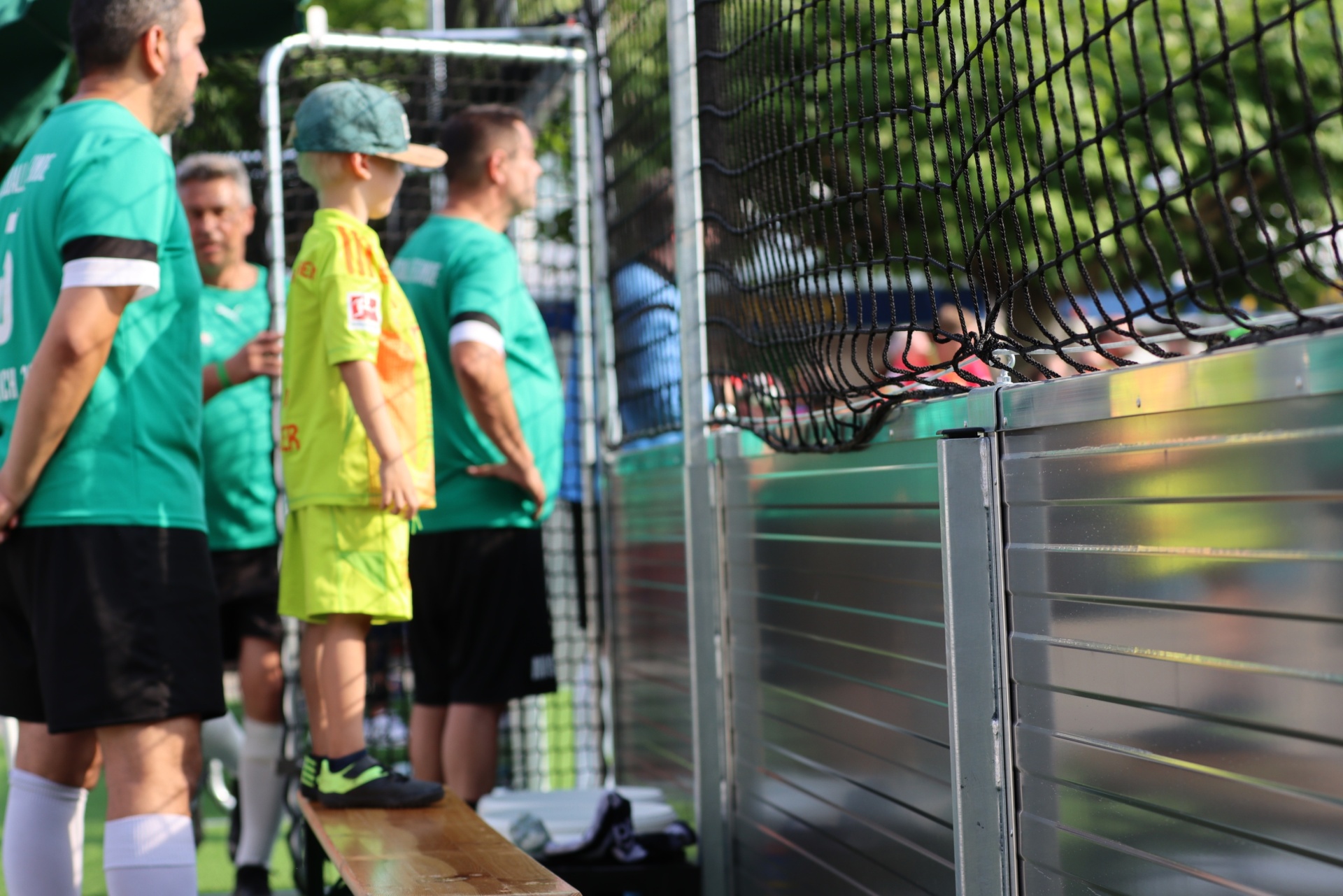 [{"clang_id":1,"value":"Fußballspieler in grünen Trikots stehen am Spielfeldrand. Ein Junge in gelbem Trikot steht auf einer Bank, Blickrichtung Spielfeld. Im Hintergrund ein Zaun."}]