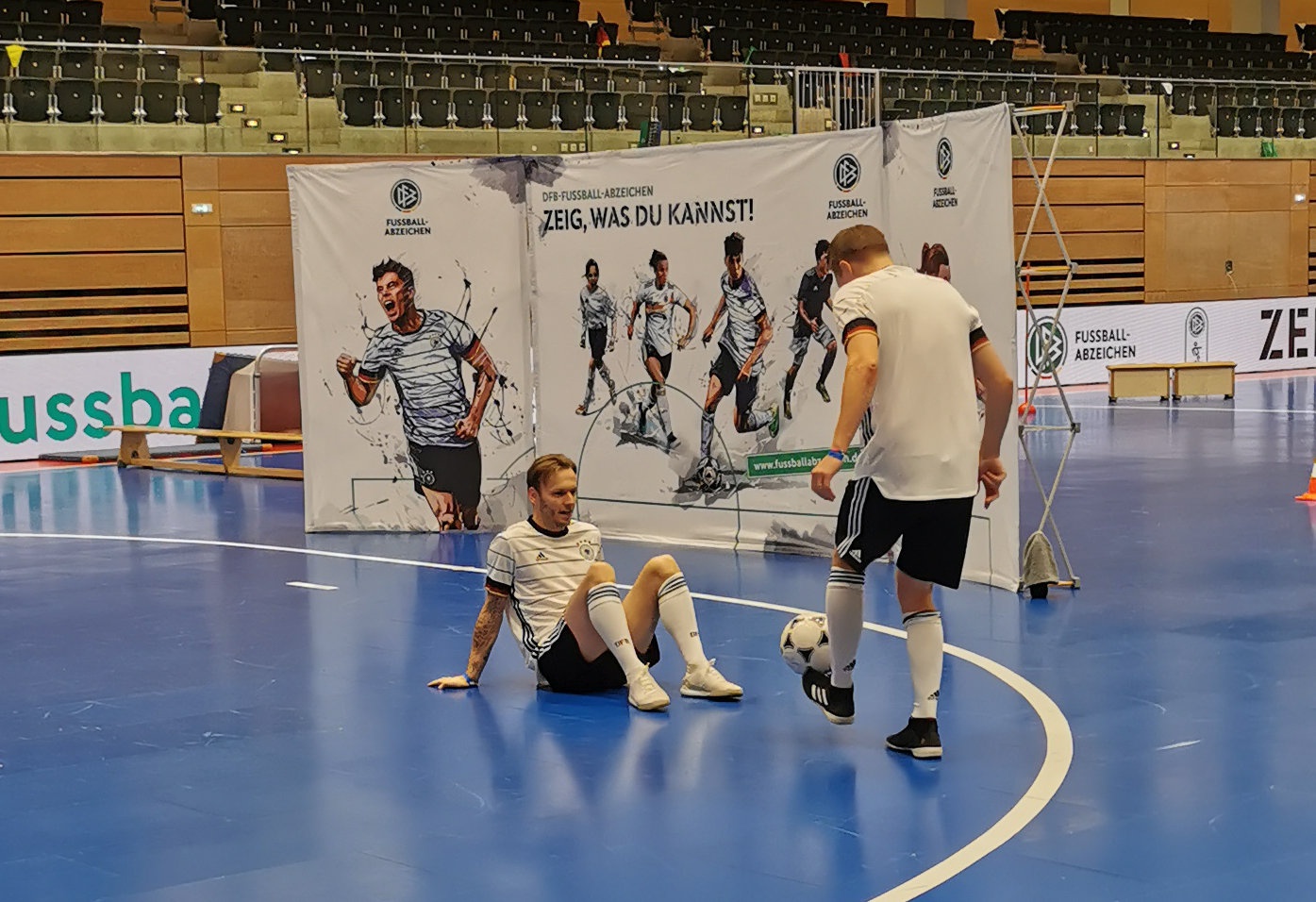 [{"clang_id":1,"value":"Drei Fußballspieler in weißen und schwarzen Trikots trainieren in einer Sporthalle. Im Hintergrund eine große Bannerwerbung mit Fußballmotiven."}]