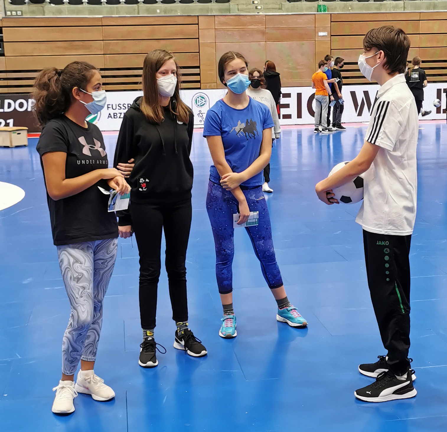 [{"clang_id":1,"value":"Fünf Jugendliche in Sporthalle. Zwei tragen Masken. Ein Jugendlicher hält eine rote Karte hoch. Blaue und graue Sportböden."}]