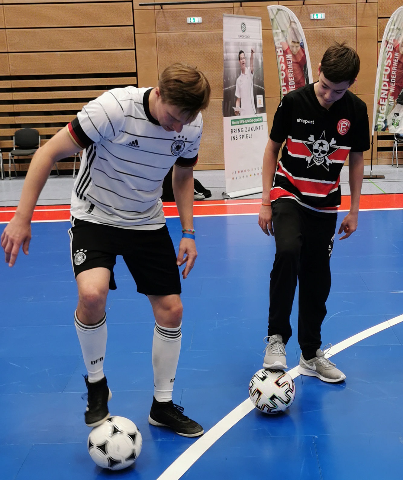 [{"clang_id":1,"value":"Zwei Jugendliche stehen in einer Sporthalle und balancieren Fußballbälle auf ihren Füßen. Links trägt ein Jugendlicher ein weiß-schwarzes Fußballtrikot, rechts einen roten Pullover mit Totenkopf-Motiv."}]