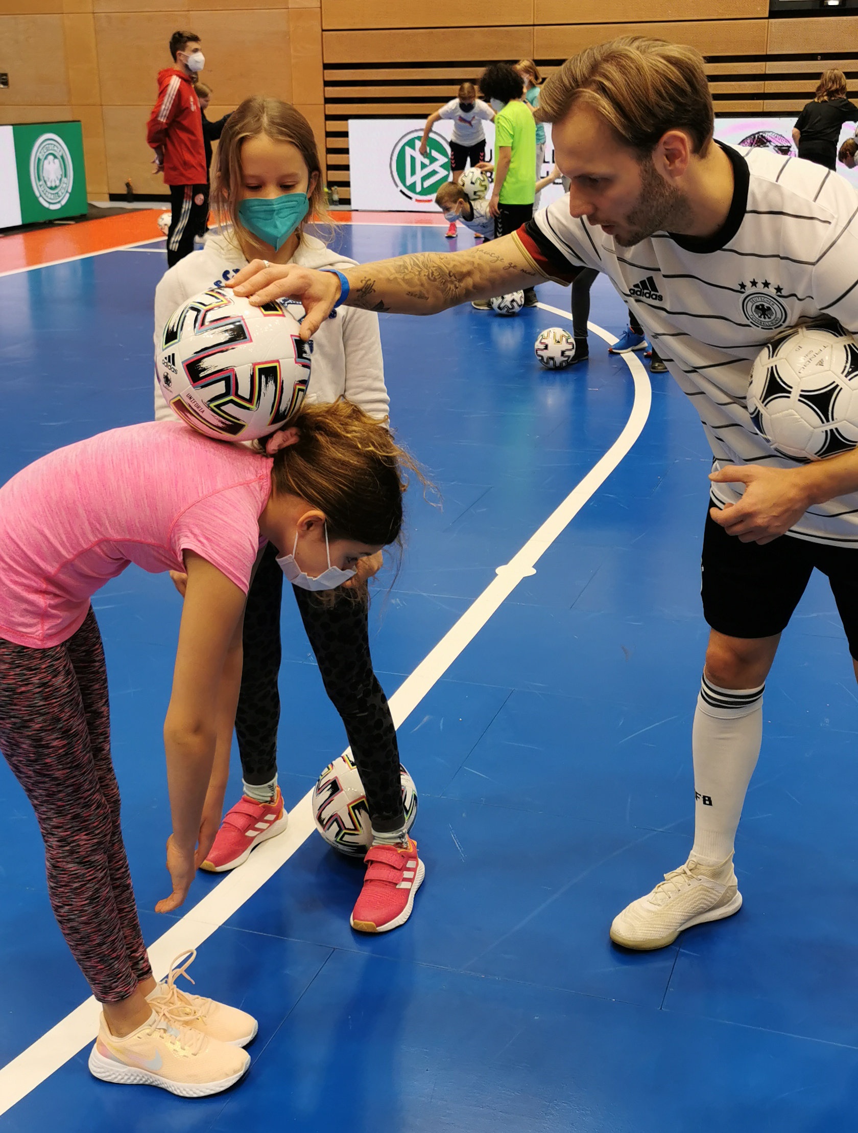 [{"clang_id":1,"value":"Fußballspieler in Deutschland-Trikot hilft zwei Mädchen beim Balancieren von Fußbällen auf dem Kopf in einer Sporthalle. Alle tragen Masken."}]