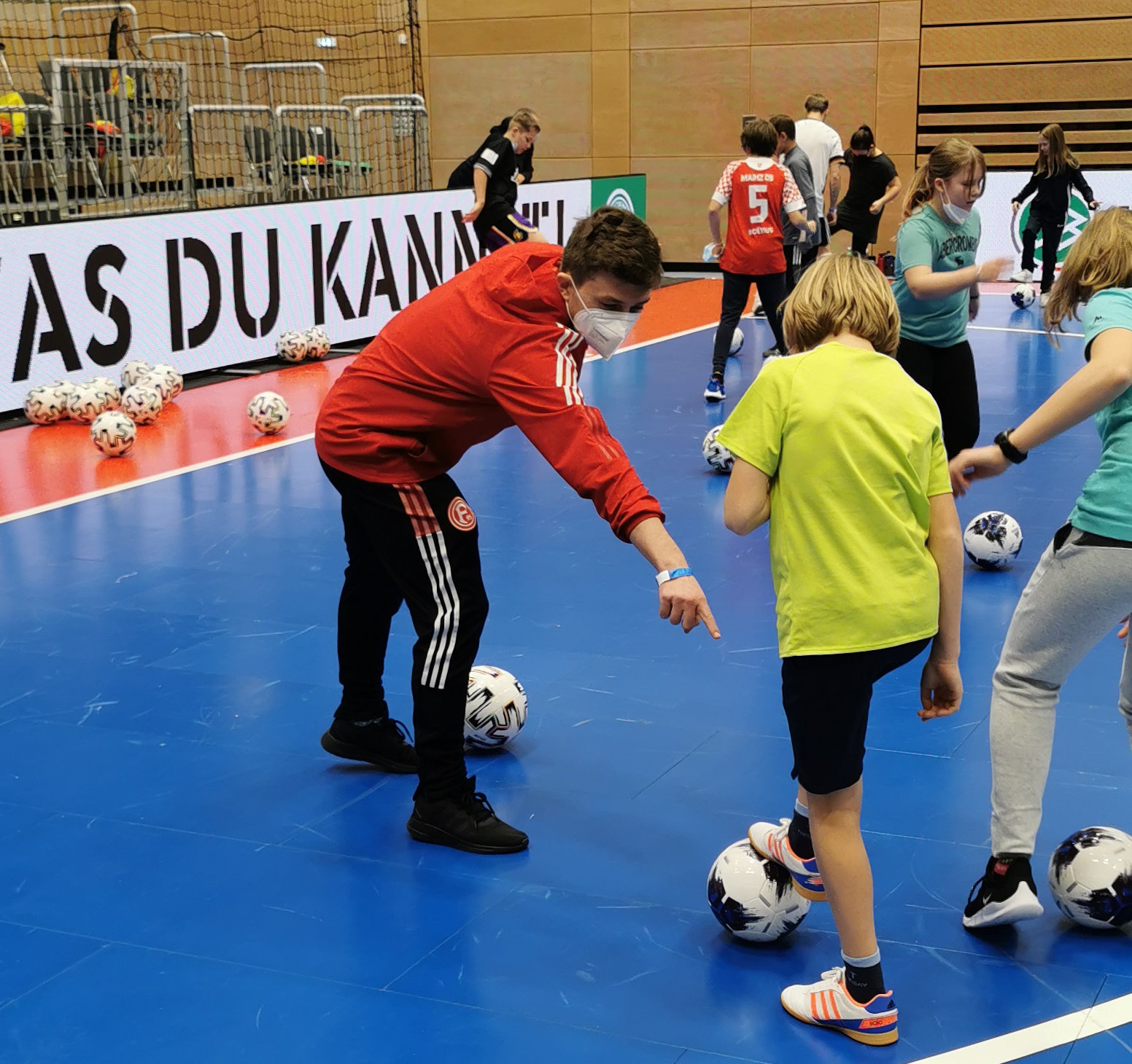 [{"clang_id":1,"value":"Helle Sporthalle. Erwachsene Person in roter Jacke und Trainingshose zeigt auf Fußballspieler-Kinder, die mit Fußbällen üben. Im Hintergrund Banner mit Schriftzug."}]